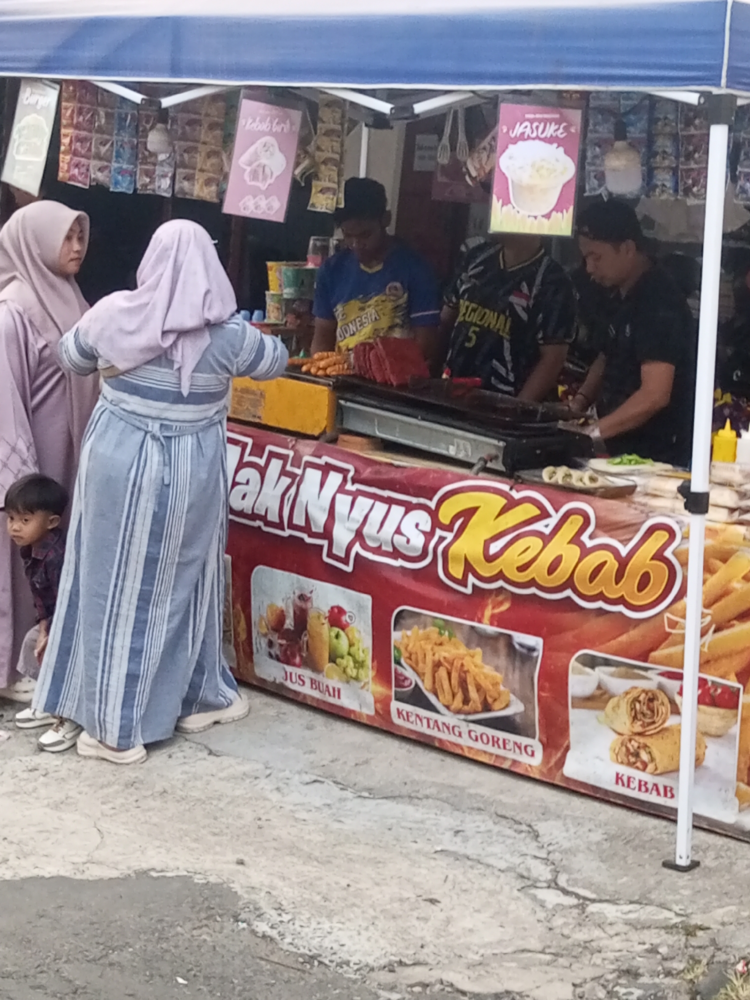 The image shows an outdoor food stall with a blue awning. In the foreground, two women wearing hijabs are visible. One woman, on the left, is wearing a light purple hijab and a light purple long-sleeved dress. The other woman, in the center, is wearing a light pink hijab and a long, blue and white striped dress. A young boy in a red and black plaid shirt is partially visible to the left of the women, looking towards the left.
Behind the women, a food stall banner displays images of food items and text in Indonesian, including "Jus Buah" (Fruit Juice), "Kentang Goreng" (Fried Potatoes), and "Kebab." Several men are working at the stall. One man, wearing a yellow and blue t-shirt with "INDONESIA" on it, appears to be cooking. Another man in a black t-shirt is also at the counter.
The background shows shelves stocked with various products, possibly snacks or ingredients, and more signage with text in Indonesian, including "Kebab Bu," and "JASUKE," which is a popular Indonesian snack. The ground is paved with rough concrete. The overall scene depicts a typical street food vendor setting in Pandansari, Indonesia.