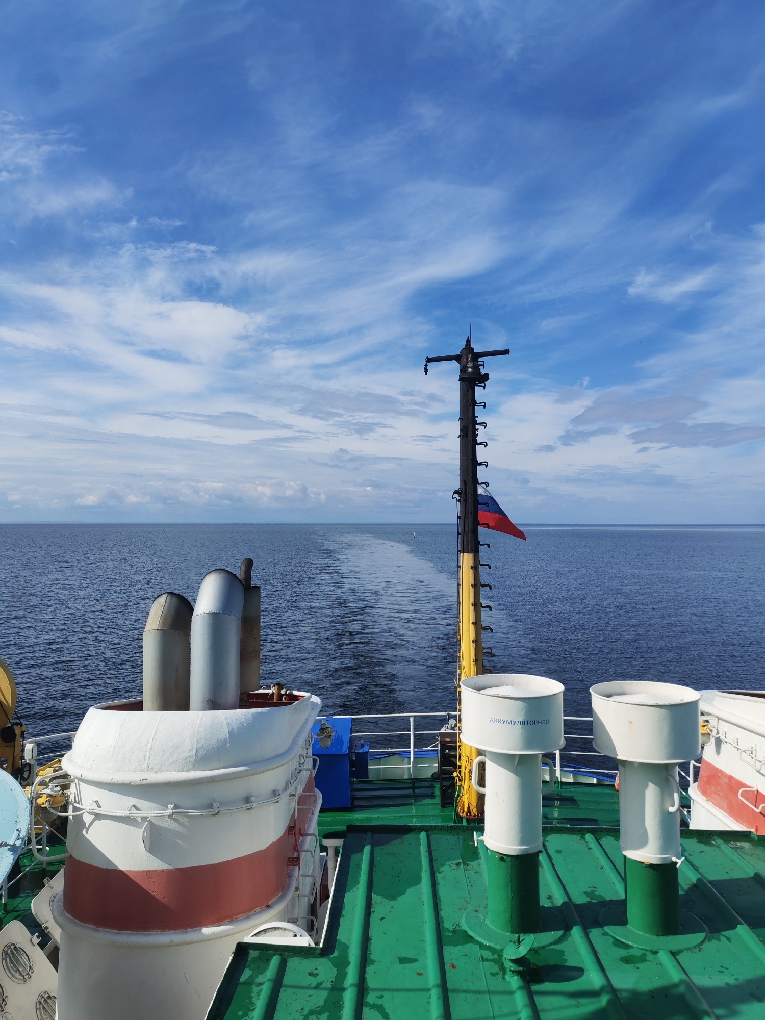 The image shows a view from the deck of a ship at sea, likely near Ozernoe Ust'e, Russia, given the context. The scene is dominated by the vast blue sea and a sky filled with scattered white clouds. In the foreground, the green metal deck of the ship is visible, featuring two white cylindrical ventilation shafts with the Cyrillic text "АККУМУЛЯТОРНАЯ" (accumulator) inscribed on one. To the left, part of a white and red life raft is visible. Further back, a tall black mast with a yellow base stands prominently, flying the Russian flag. The ship is creating a wake in the water, indicating it is moving forward. The overall environment is maritime and industrial. There are no people visible in the frame.