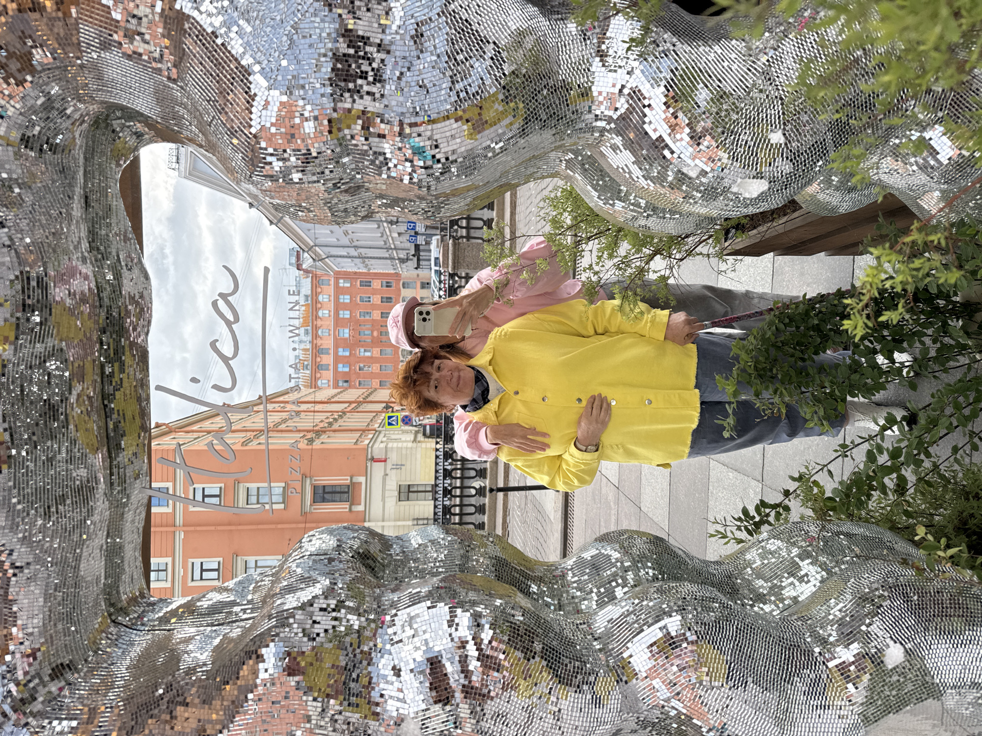 Two women pose for a selfie near a mirrored sculpture in Saint Petersburg, Russia. One woman wears a yellow jacket and the other wears a pink jacket and hat. The sculpture reflects the buildings and sky of the city. Foliage is visible to the right of the sculpture.