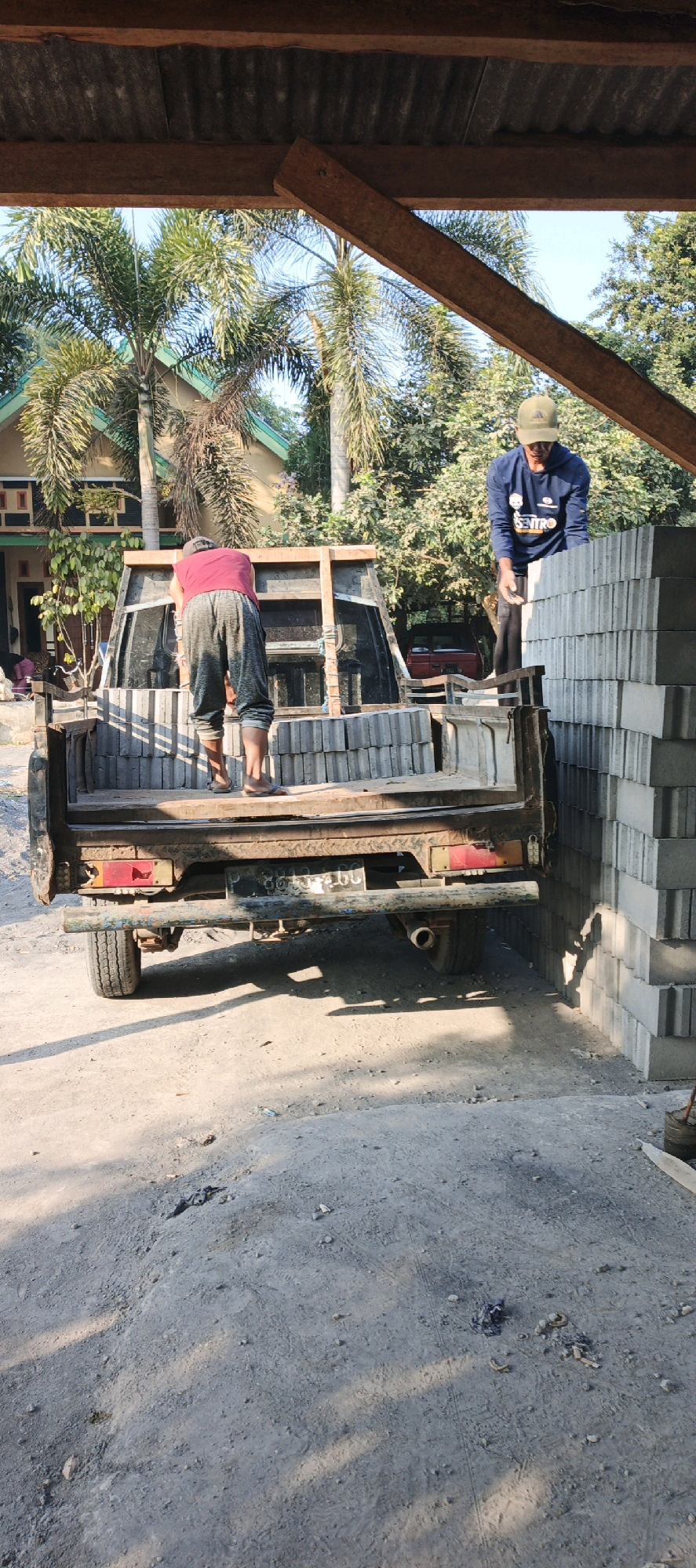 In Kasiyan, Indonesia, two men are seen working outdoors under a covered structure, likely a shed or open-air workshop, with palm trees and a house visible in the background. The ground is unpaved and dusty, with piles of gray concrete blocks stacked high to the right of the frame.
In the foreground, a rusty, dark-colored pickup truck is partially visible from the rear. The truck bed is loaded with more concrete blocks, some stacked neatly and others appearing haphazardly placed. A wooden frame structure has been erected in the truck bed, possibly for transporting or securing the load.
One man, wearing a red t-shirt and gray sweatpants, is bent over in the truck bed, appearing to organize or unload the blocks. He is barefoot. To his right, another man, wearing a dark blue long-sleeved shirt and a green cap, is standing on the edge of the truck bed and a stack of blocks. He is holding a gray concrete block, seemingly in the process of either placing it or receiving it.
The lighting suggests it is daytime, with shadows cast by the structure and the truck onto the ground. The overall scene depicts manual labor related to construction or material handling in an outdoor, possibly rural, setting.