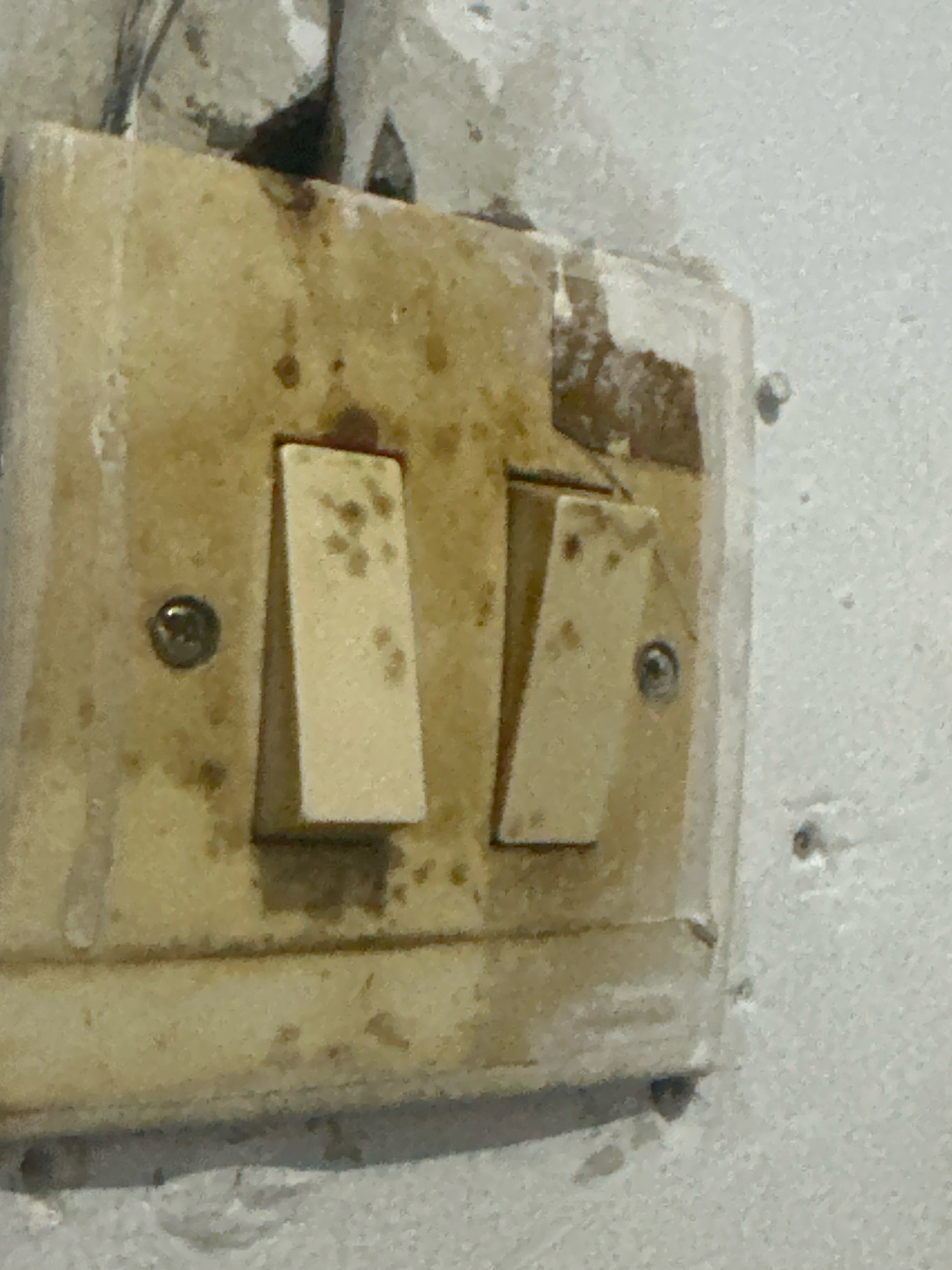 In Margamulya, Indonesia, a close-up, eye-level shot captures a dirty, yellowing light switch plate affixed to a textured white wall. The plate, showing signs of age and grime, is mounted with two silver screws. Two light switches, each a cream-colored rectangular toggle, protrude from the plate. The left switch is angled downwards, while the right switch is angled upwards. The wall surrounding the switch plate is rough and uneven, with some areas appearing chipped or damaged. The lighting is dim, casting subtle shadows and highlighting the textured surface of the wall and the discolored state of the switch plate.