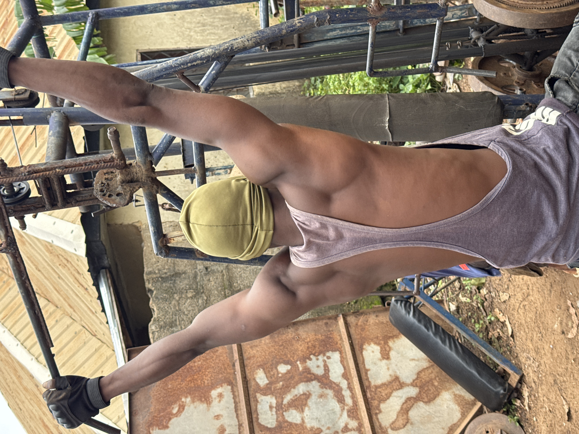 In Akwanga, Nigeria, a dark-skinned man wearing a green durag and a grey tank top is performing a lat pulldown exercise on a rusty, homemade weight machine. He is gripping a metal bar with gloved hands, his arms extended upwards. The machine is outdoors, with a corrugated metal roof structure in the background. In the foreground, a black padded bench is visible, along with loose dirt and fallen leaves. The scene suggests a utilitarian workout environment.