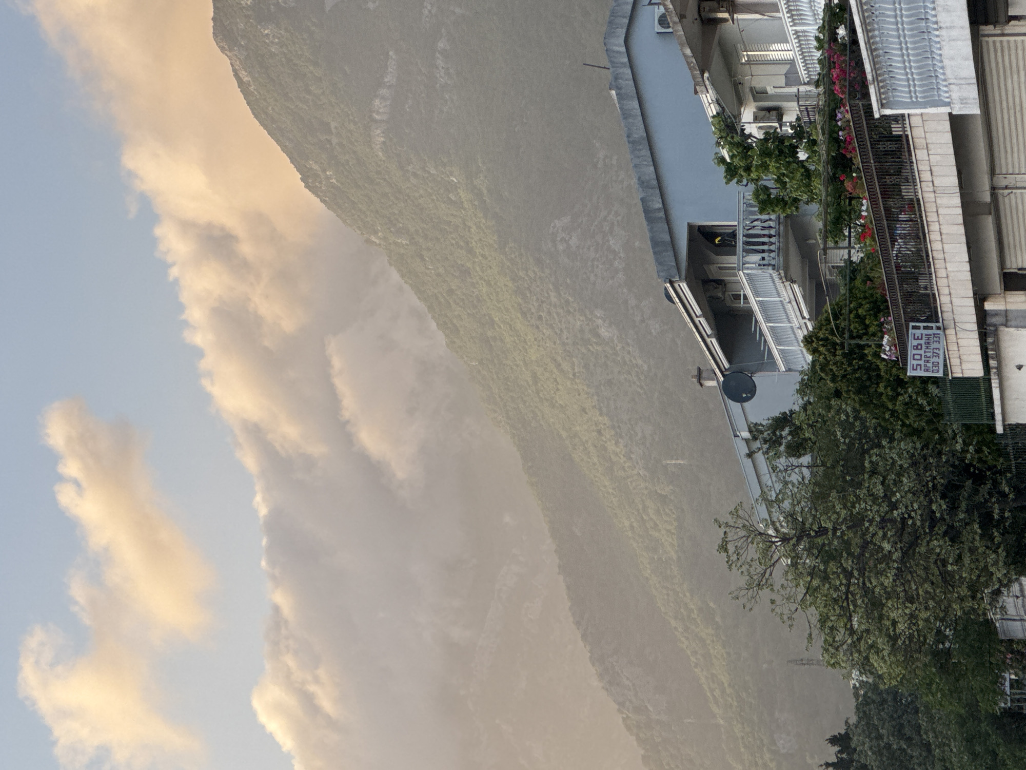 The image shows a view in Sutomore, Montenegro. In the background, a large, green, mountainous landscape is partially obscured by clouds. To the right, a multi-story building is visible, featuring balconies adorned with flowers and plants. Signs and an antenna are attached to the building. Green trees surround the building. The sky is light blue with some white and orange clouds.