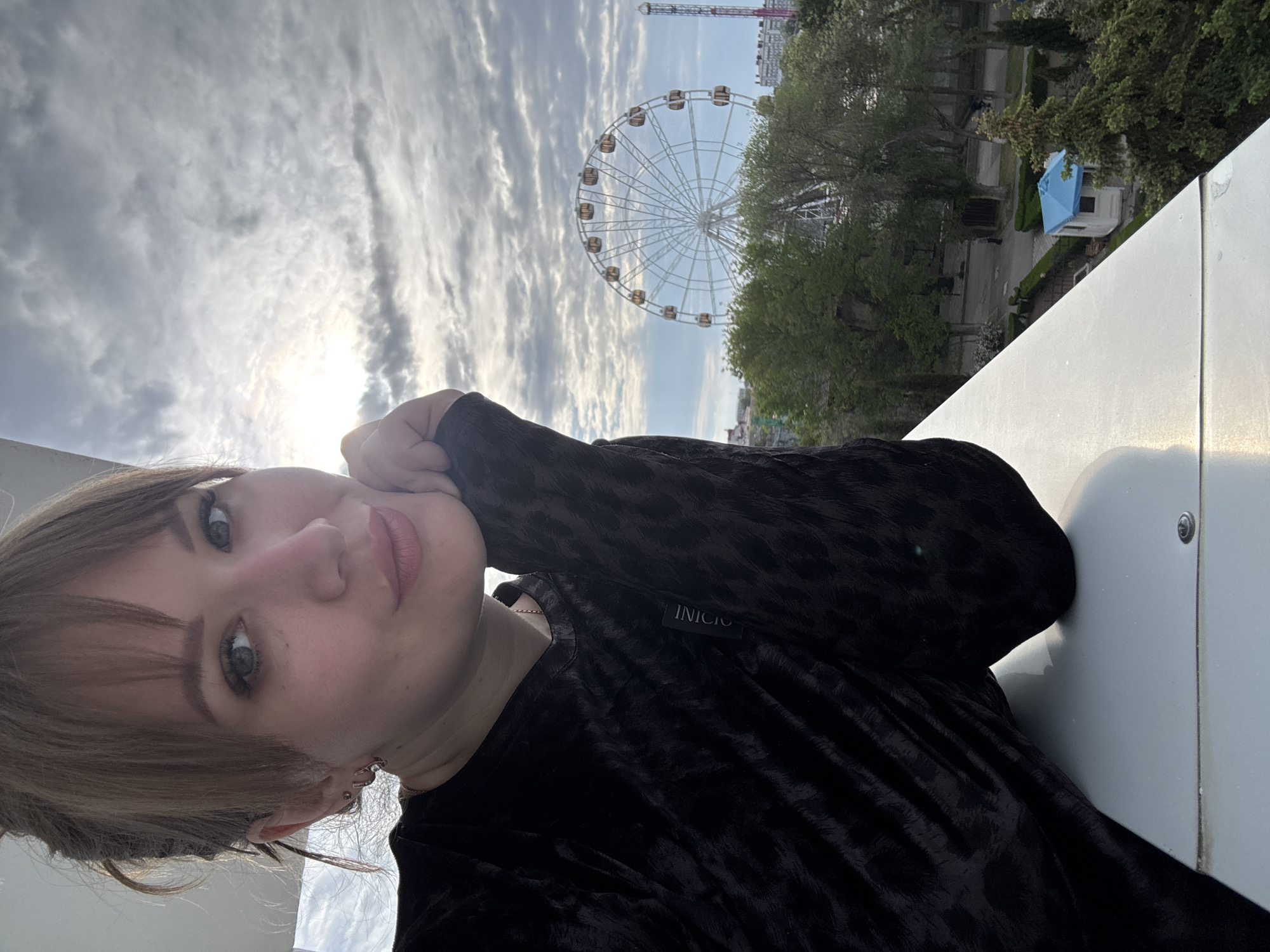 A woman is posing for a selfie against the backdrop of the Yevpatoriya Ferris Wheel, located in Yevpatoriya, Crimea, Ukraine. The Ferris wheel is centrally framed with wispy clouds above. Green trees flank it, and a blue-roofed structure is visible beneath the trees to the right of the wheel. The woman is on an elevated structure, indicated by a white railing. She has her cheek resting on her hand, wearing a dark-colored velvety top with the word "INICIO" visible on a tag. Her light brown hair is styled with bangs. The sky is overcast with the sun partially visible.