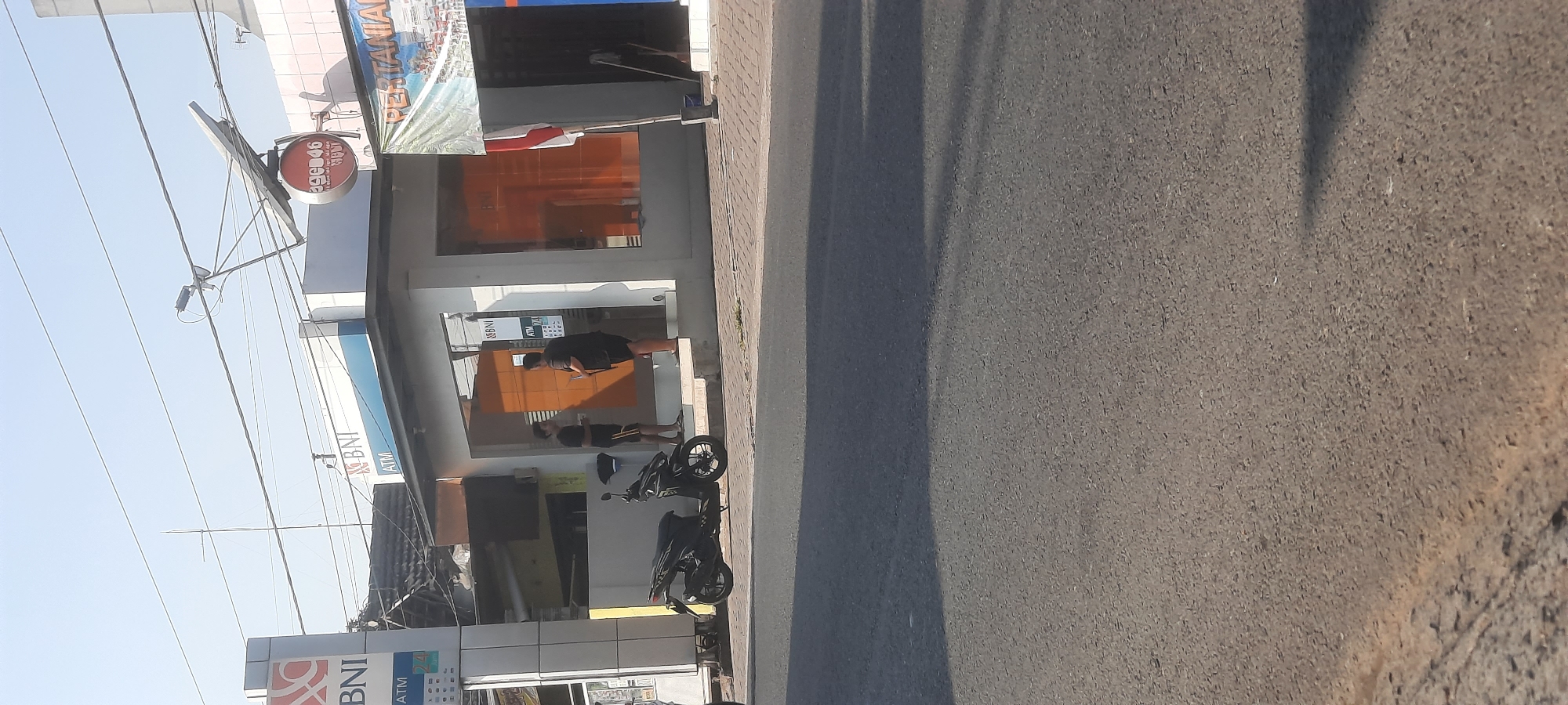 The image shows a street scene in Selopuro, Indonesia, with a focus on a BNI (Bank Negara Indonesia) ATM and office building. Two individuals, both wearing dark clothing, are standing outside the building, one near the entrance and the other further in. A black motorcycle is parked on the sidewalk to the left of the ATM. Above the ATM, a satellite dish is mounted on a pole. A banner with the word "PERTANIAN" is visible on the right side of the building. The street in the foreground is paved and appears to be a two-lane road with a sidewalk. The sky is clear and blue, suggesting a sunny day.