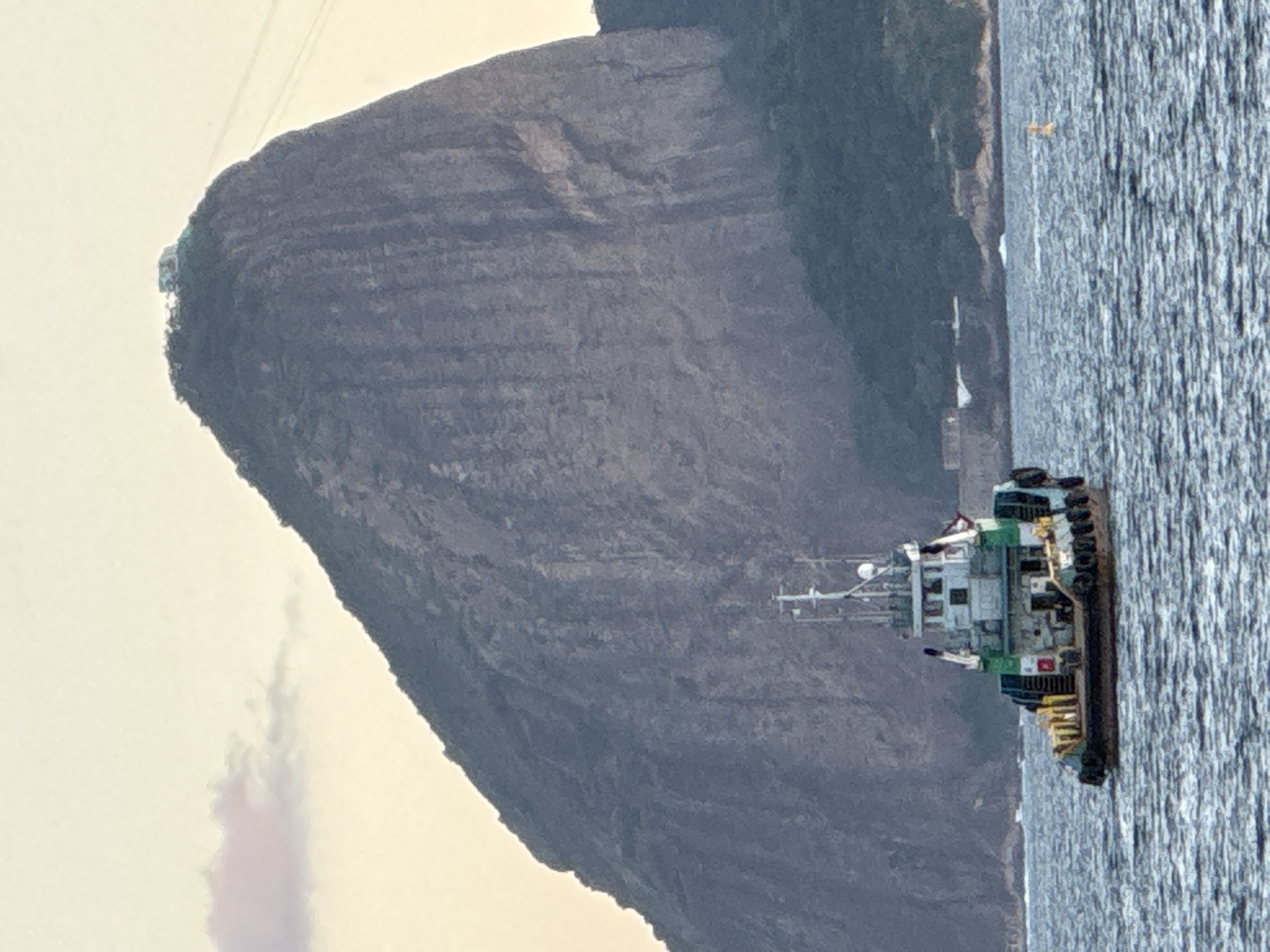 The image depicts a tugboat sailing on a body of water in front of Sugarloaf Mountain, a prominent landmark located in Rio de Janeiro, Brazil, rather than Niterói. The mountain, characterized by its steep, rocky face, dominates the background with a structure visible at its summit and what appears to be a cable car system ascending its side. The sky is a pale, hazy yellow, suggesting either dawn or dusk. The water is choppy, and the tugboat is positioned in the foreground, showing its deck with various structures, railings, and tires along its hull. In the distance, along the shoreline, some buildings and dense vegetation are visible at the base of smaller hills. A yellow buoy is also visible in the water to the right of the tugboat.