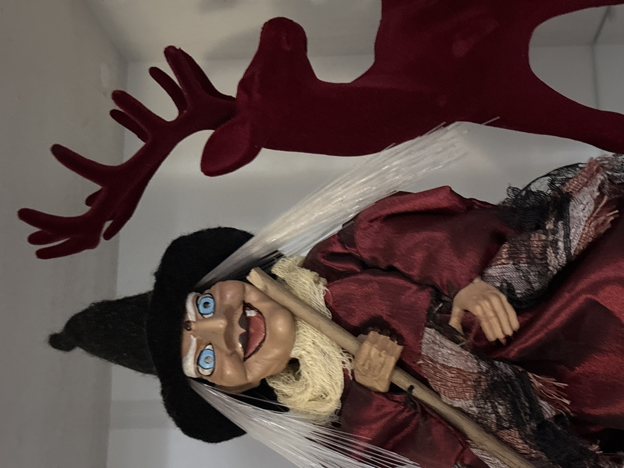 The image shows a close-up of a witch doll and a red reindeer decoration. The witch doll is on the left, wearing a black pointed hat, a red robe, and a cream-colored scarf. She has white hair, blue eyes, and a wide smile with sharp teeth. She is holding a wooden broomstick. To the right of the witch doll is a red, flat cutout of a reindeer with antlers. The background is a plain, light-colored wall. There is no indication of the location being Chaikovsky, Russia.