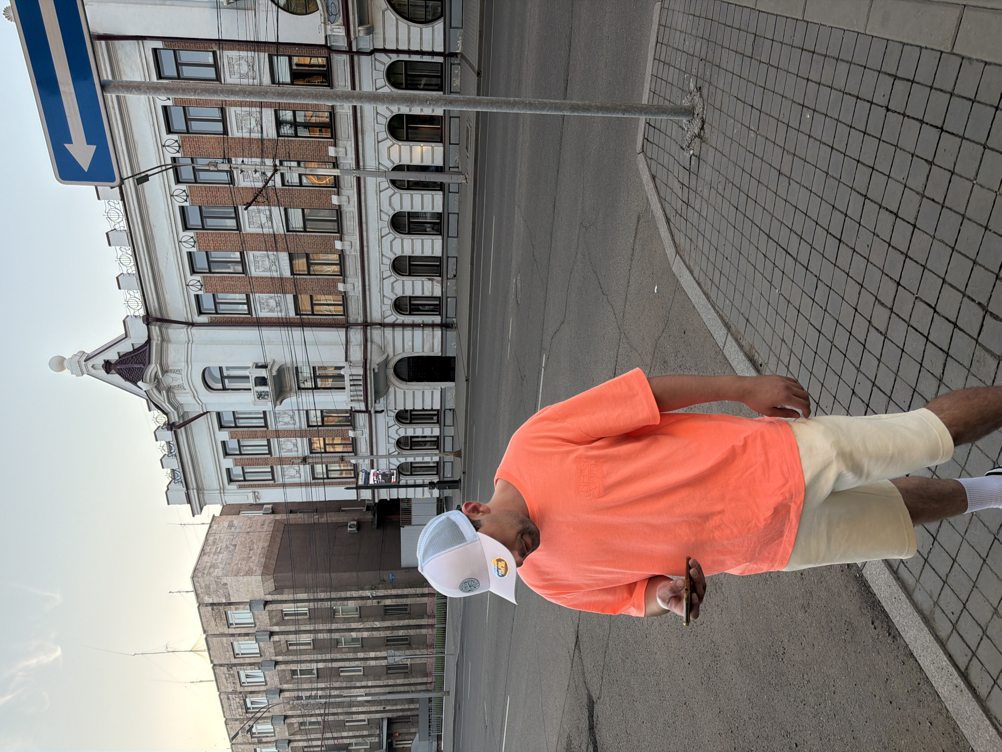 This eye-level outdoor shot captures a person standing on a paved sidewalk in a city, likely Krasnoyarsk, Russia. The main subject is a male, dressed in a vibrant orange short-sleeved t-shirt, light cream-colored shorts, white socks, and white sneakers. He wears a light-colored mesh baseball cap with a circular logo on the front and holds a smartphone in his right hand, looking downwards or to the side.
Behind the person, to the left, stands a multi-story building characterized by ornate white stucco details, red brick sections, and numerous windows, some arched. The building features decorative cornices and a prominent peaked roof section on its far left. In the background, across the street to the right, another multi-story building with a more modern, textured facade and multiple windows is visible.
The person stands on a sidewalk made of square gray pavers, adjacent to an asphalt road with white lane markings. A blue rectangular street sign featuring a white downward arrow is mounted on a metal pole, positioned above and slightly behind the person. The sky is clear, suggesting fair weather, and the soft, diffused lighting indicates late afternoon or early evening.
Visible text includes a logo on the person's cap and stylized text on his t-shirt that appears to read "NEON" above another word.