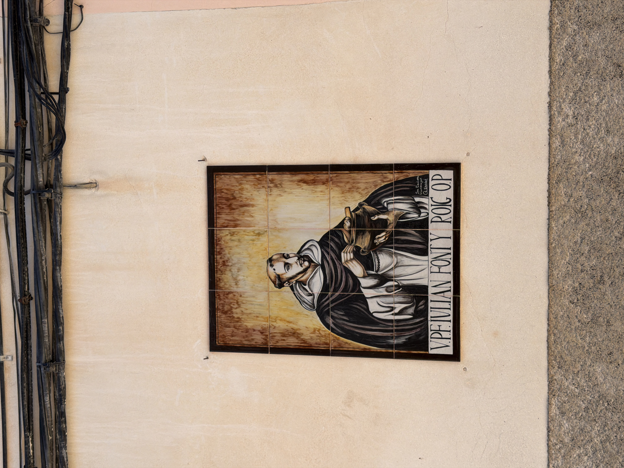 A tiled mural depicting a Franciscan friar holding a pot adorns a stucco wall on a street in Manacor, Spain. The friar, identified by the inscription "V. PE. JULIAN FONTY ROIC OP" below the image, is depicted with a tonsured head, beard, and a brown scapular over a white habit. The background of the mural is a textured blend of brown and yellow, and a small signature reads "Sa Taulera Llucmajor @Saase". The mural is framed by a dark border and mounted on a pale beige wall. Above the mural, electrical cables are strung across the top of the frame. The bottom of the image shows a rough-textured grey stone base along the wall.