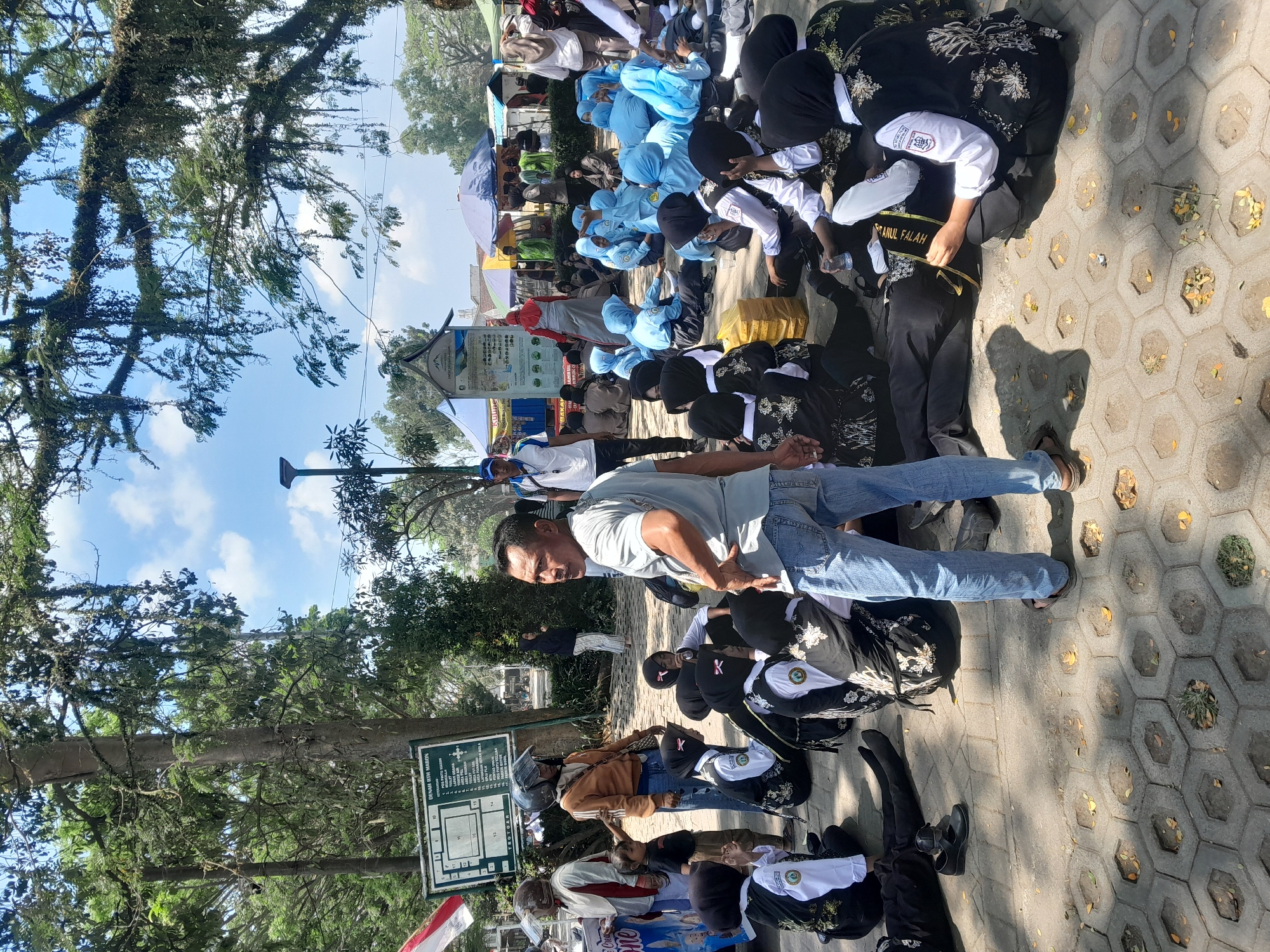 A man in a grey t-shirt and blue jeans stands in the foreground, facing away from the camera, speaking to a group of young people sitting on the ground. The group includes several individuals wearing black and white school uniforms with headscarves. They appear to be listening to the man.
In the background, there are several tents and stalls, suggesting an outdoor event or market. Some people are seen walking around the area, including a person wearing a blue shirt and a cap, and another individual in a red headscarf and grey skirt. Trees provide shade over the scene.
The ground is paved with hexagonal tiles. A sign with text and a map is visible on the left side of the image, partially obscured by trees. The overall scene suggests a lively gathering in an outdoor public space in Genteng, Indonesia.