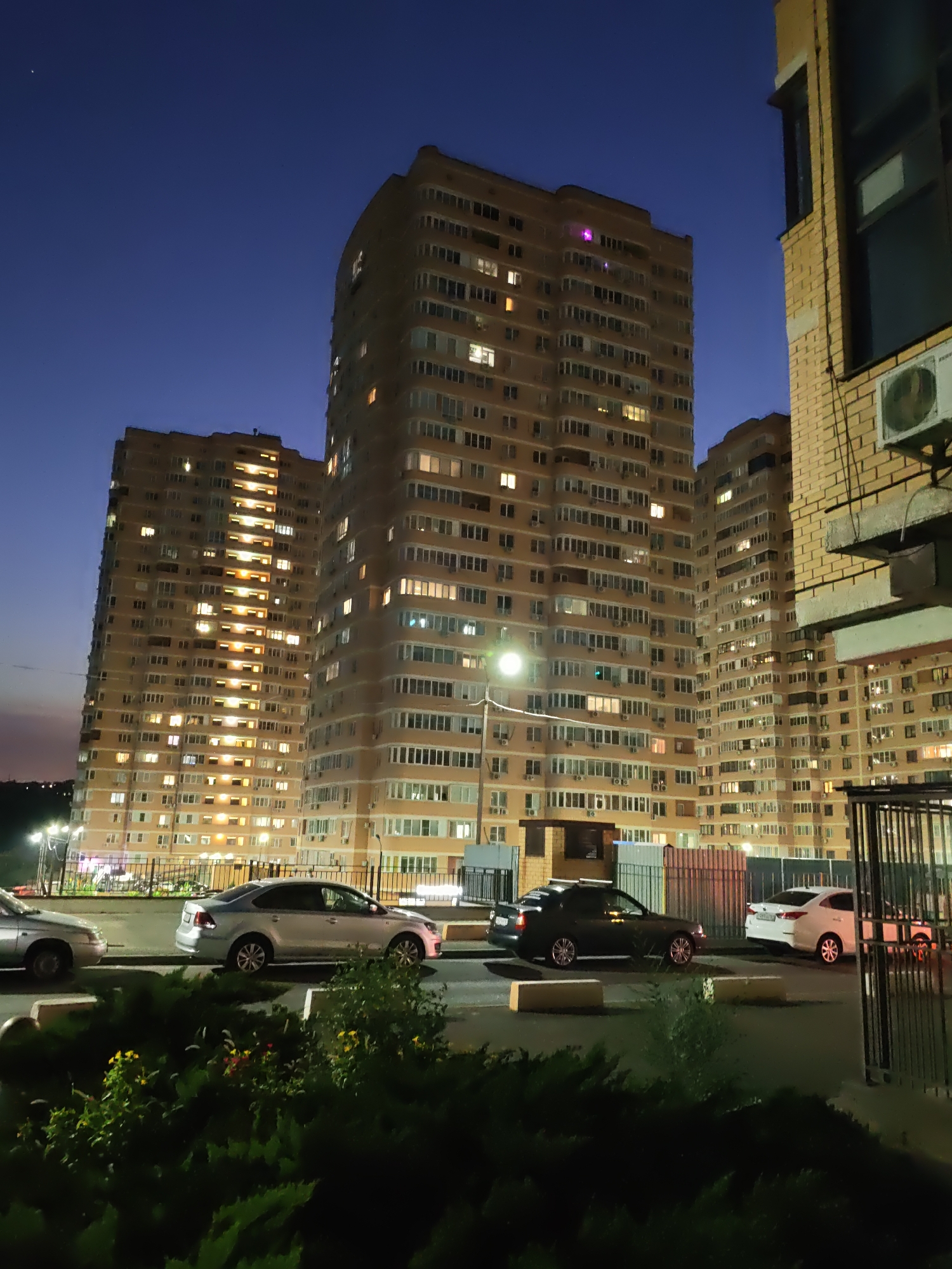 The image depicts a nighttime scene in a residential area of Rostov-on-Don, Russia, featuring tall apartment buildings and parked cars. The buildings, illuminated by numerous lit windows, dominate the background. In the foreground, several cars are parked on a paved surface. Lush green bushes with some flowering plants are visible in the immediate foreground. A metal fence is present in the middle ground. The sky is dark blue, indicating it is twilight or early night. No people are visible in the image.