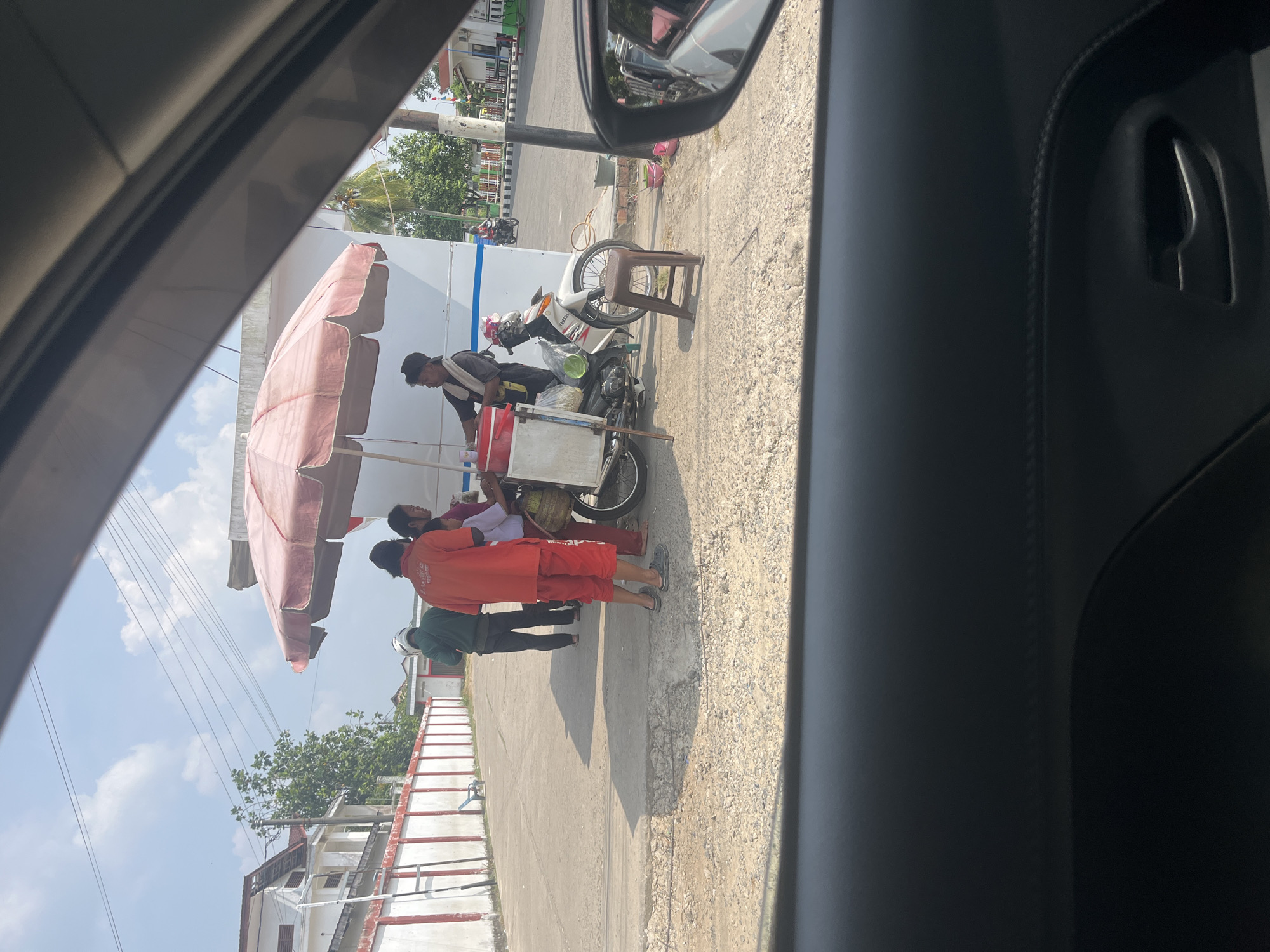 The image, taken from inside a vehicle, shows a street scene in Sekayu, Indonesia. A vendor, wearing a red shirt and red pants, stands under a large, faded pink umbrella. The vendor is interacting with at least two other people, one of whom appears to be a child wearing a white shirt. A motorbike with a sidecar is parked next to them. The sidecar is filled with goods, possibly food items, and has a basket attached. A small, brown plastic stool is placed near the motorbike. In the background, there is a white building with a red trim and a fence. Trees are visible behind the building. The ground is paved with concrete and gravel. The sky is blue with scattered clouds. The image appears to have been taken during the day, likely in the morning or afternoon due to the sunlight and shadows.