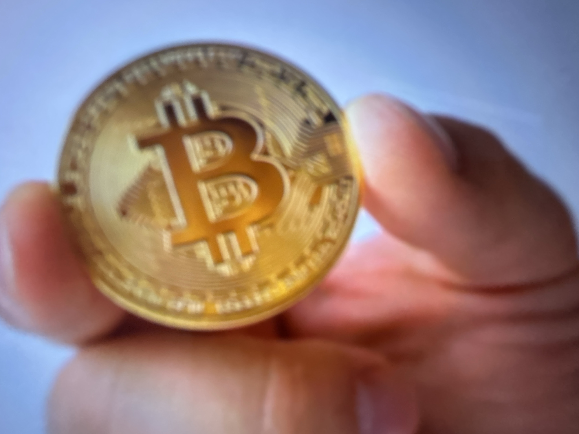 A person's hand, with visible skin texture and fingernails, is holding a gold-colored Bitcoin coin. The coin is partially in focus, showing the characteristic "B" symbol. The background is blurred and appears to be a light blue sky. The scene is not located in Gelendzhik, Russia, or any specific city.