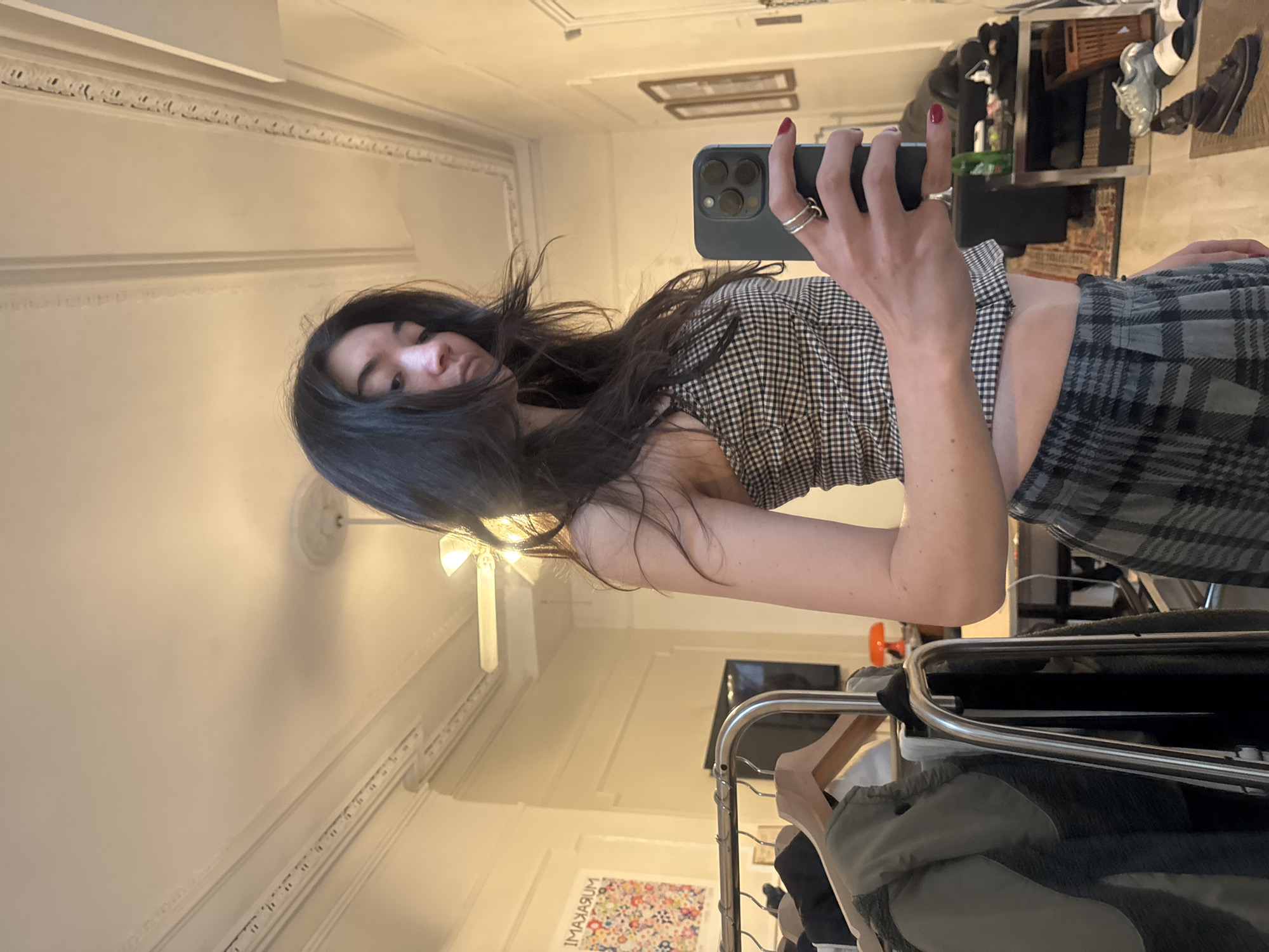 A woman is taking a selfie in a room that appears to be in a New York City apartment. She is holding a smartphone in her right hand, showing the camera towards herself. She has long, dark hair. She wears a black and white checkered top and a gray and black plaid skirt. The room is decorated with ornate moldings, indicative of older buildings in NYC. A clothing rack with garments is visible. In the background, there is a glimpse of a framed artwork on the wall.