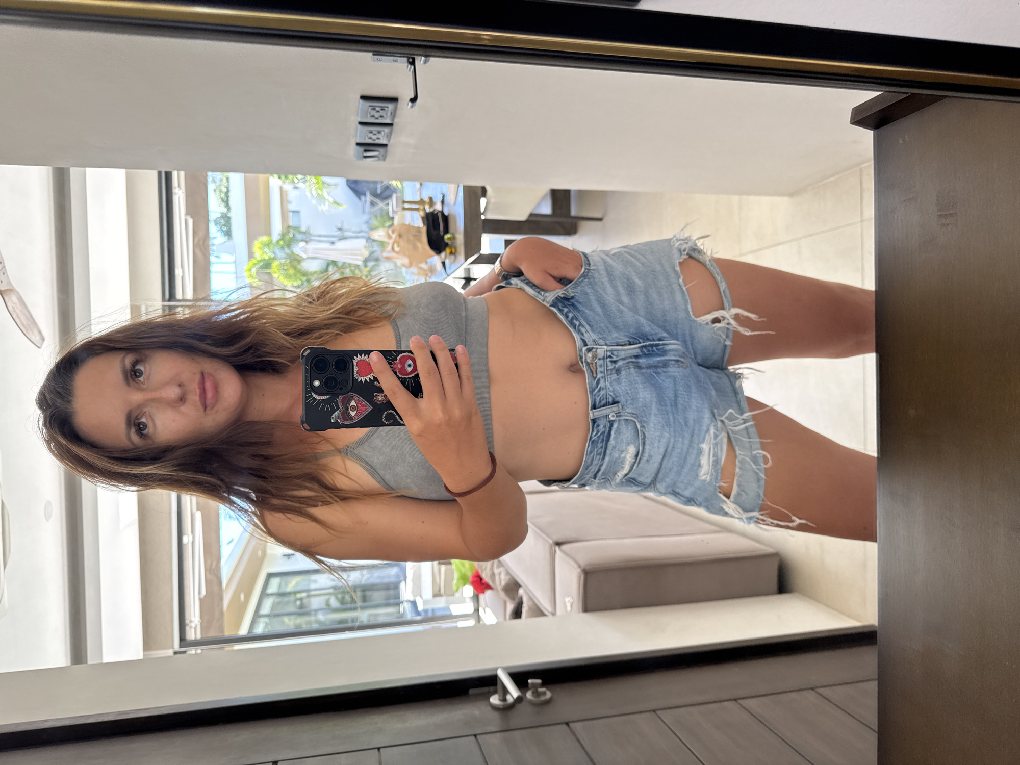 A woman is taking a selfie in a mirror. She is wearing a grey crop top and ripped denim shorts. She has long, wavy brown hair and is holding her phone in her right hand. The background shows a bright, airy room with a ceiling fan, a couch, and a table with chairs. The setting appears to be a modern home or hotel.