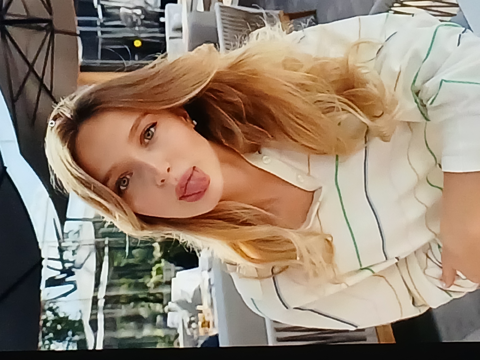 A young woman with blonde hair is sticking her tongue out at the camera. She is wearing a white shirt with horizontal green and brown stripes. The background suggests an outdoor seating area, possibly a restaurant or cafe, with tables and chairs visible. The lighting is bright, indicating daytime. There are no clear indicators in the image to determine the specific location within Dnipro, Ukraine.
