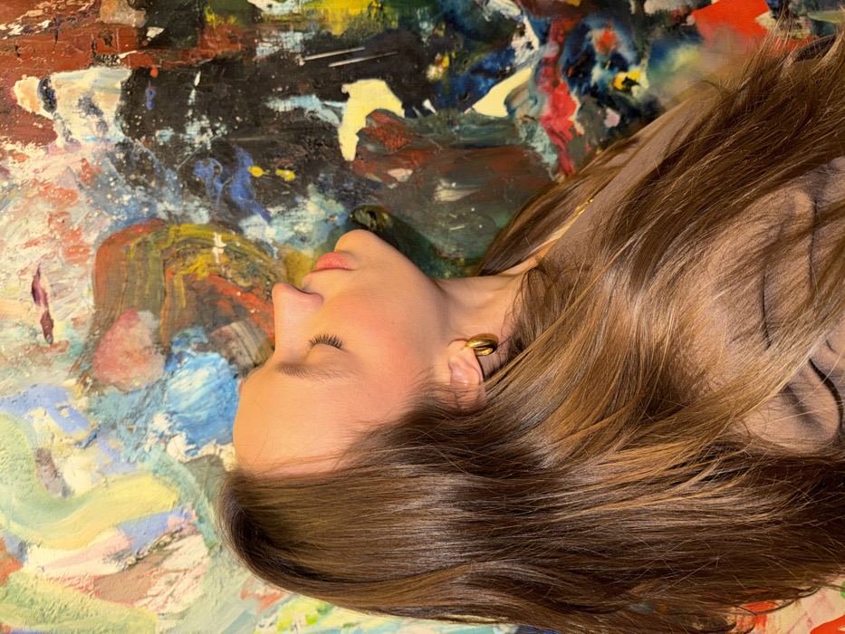 A single individual, appearing to be a young adult female, is depicted in profile, facing left, with her head tilted upwards and slightly backward. Her eyes are closed and her mouth is slightly open. She has long, straight, light brown hair flowing over her shoulders and chest. A single gold-colored hoop earring is visible on her right ear, and she wears a thin gold-colored chain around her neck, partially obscured by a light brown or beige garment. The subject is positioned against an abstract painting, which serves as the background. This artwork features a diverse and vibrant palette of colors including blues, greens, yellows, reds, oranges, blacks, and whites, applied with thick, impasto brushstrokes creating a textured surface. The scene takes place indoors. No specific visual cues are present in the image to identify a precise location within the city of Budva, Montenegro.