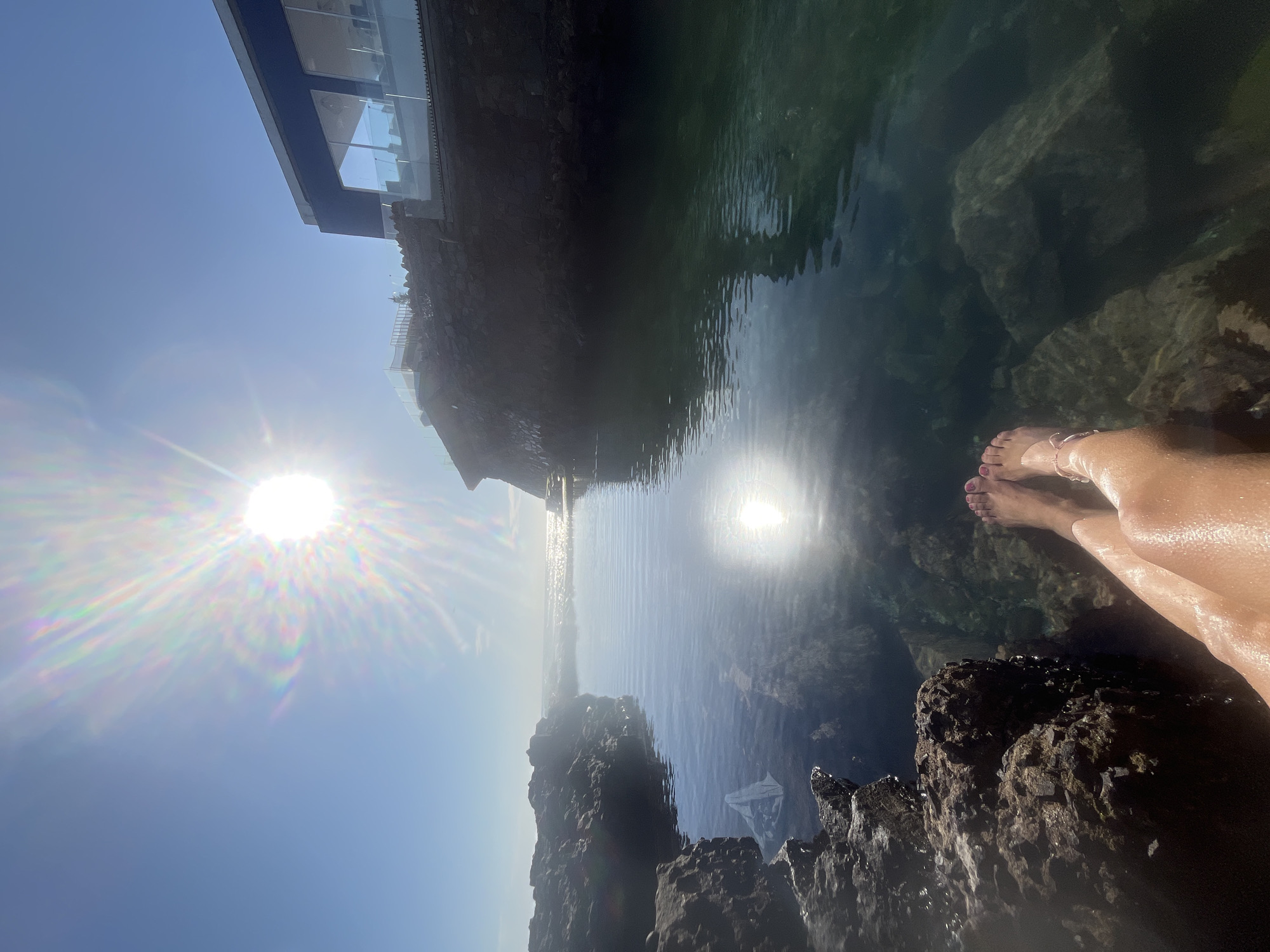 A high-angle, vertically oriented, extreme wide shot depicts a sunny seaside scene in an unknown location. A structure with glass railings and a rocky stone base sits on the upper left. Below it, the water's surface reflects the sun. The water is surrounded by dark rocks. A pair of legs are positioned on the upper right with pink toenails and strappy sandals. The sky is visible on the left, with the sun as a bright focal point.