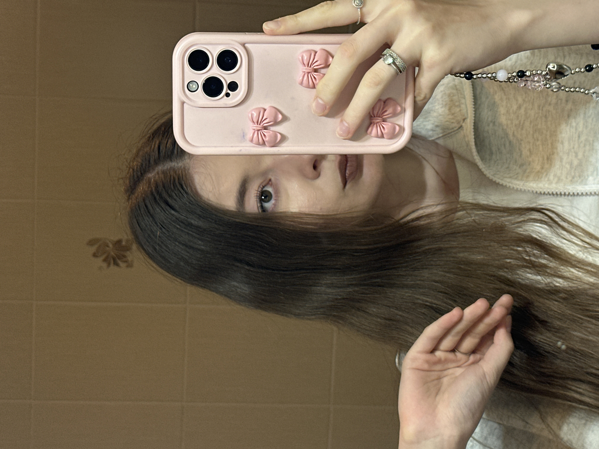 A person is holding a pink smartphone with a pink case decorated with three-dimensional pink bows in front of their face in a mirror. The phone partially obscures the face, but one eye and the mouth are visible. The person has light skin, brown hair, and is wearing rings on their fingers. They also wear a light gray zipped top. A beaded phone charm is hanging on their hand. The reflection shows a bathroom with light brown tiled walls. The exact location in Kazan, Russia, is not discernible from the image.