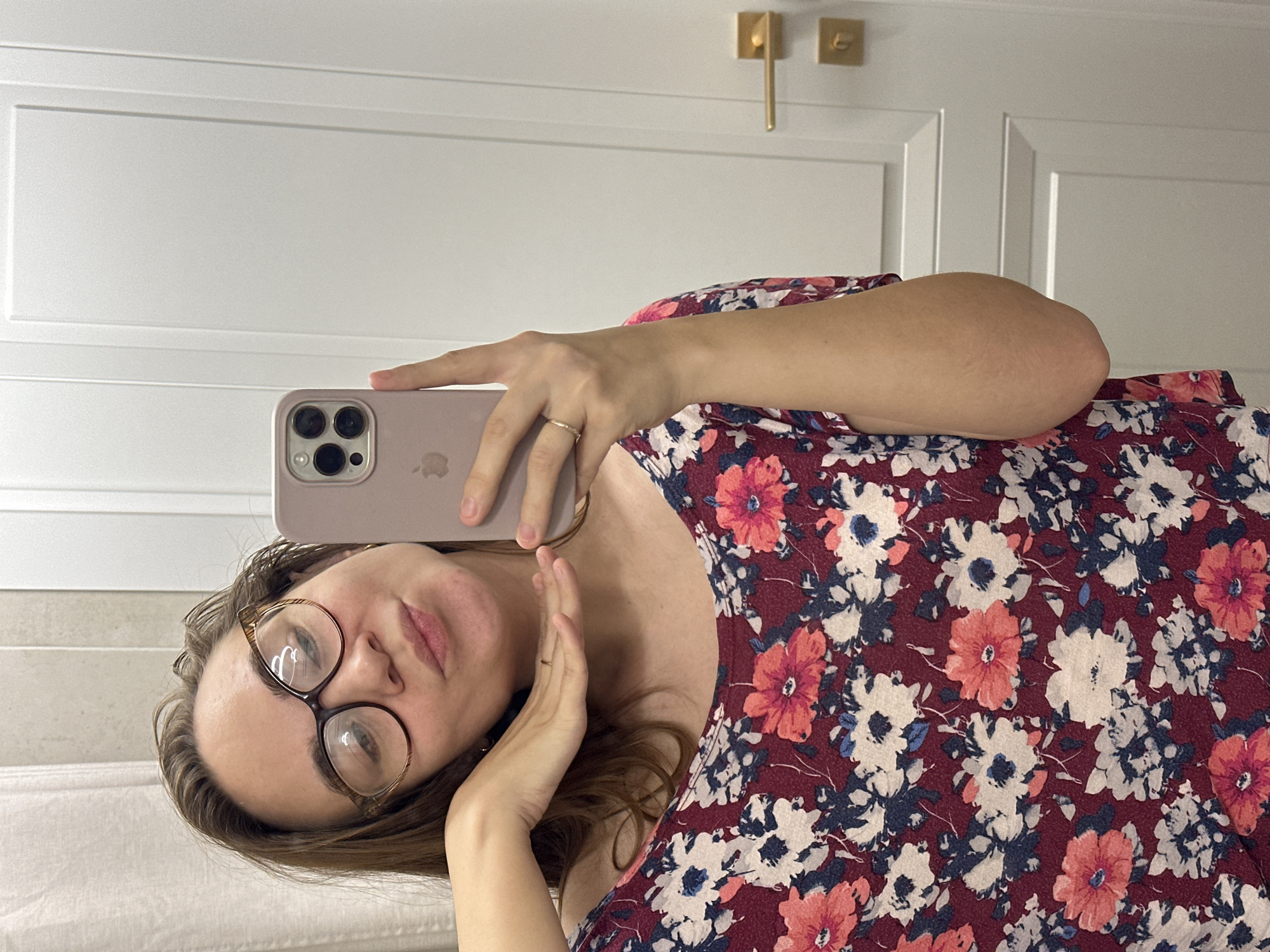 A fair-skinned woman wearing glasses and a maroon floral-print dress is holding a rose-colored smartphone. The background shows a white paneled wall with gold hardware. The image does not contain information about the location in the city of Kharnavy, Russia.