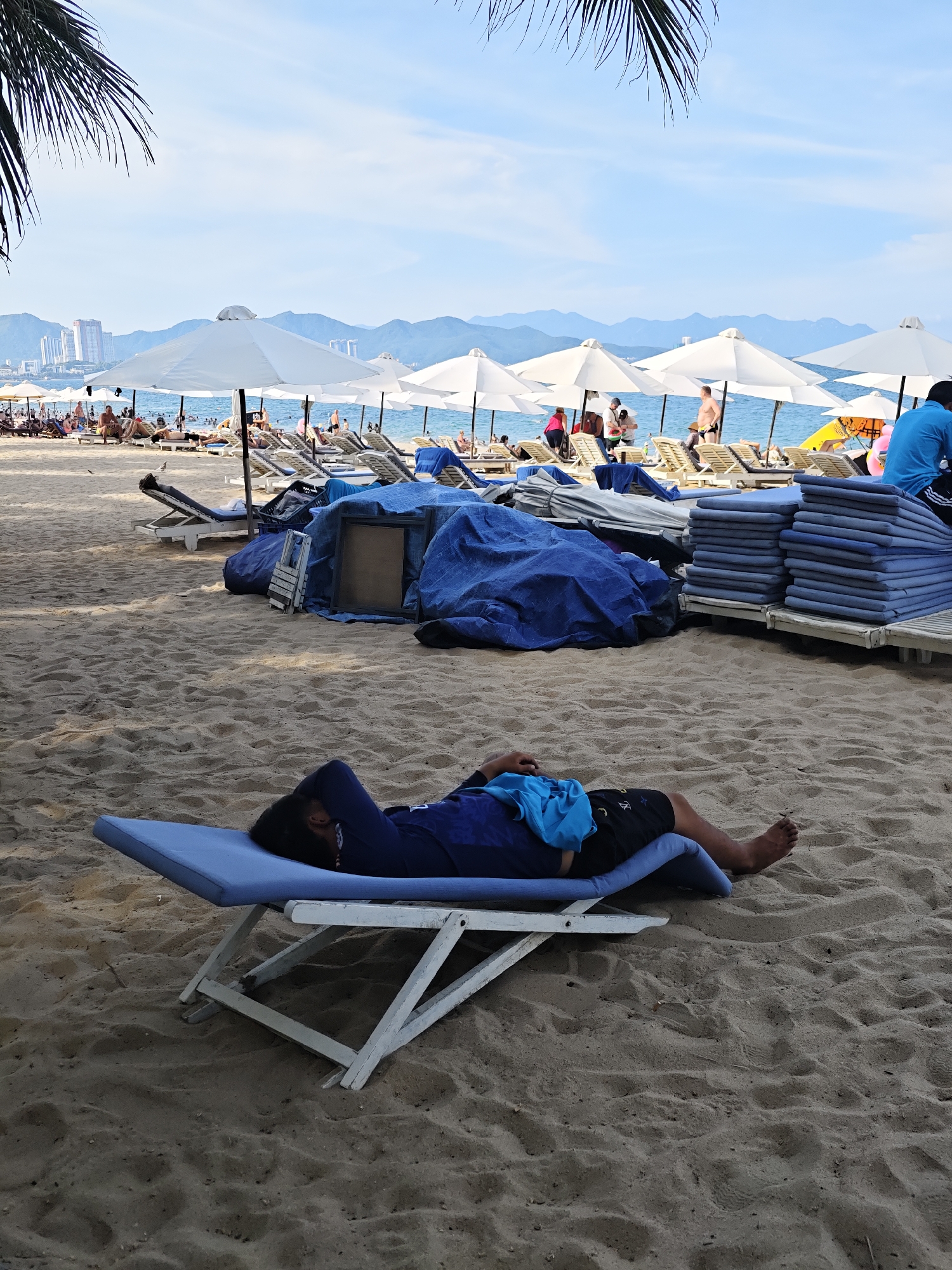 The image depicts a beach scene in Nha Trang Ward, Vietnam. In the foreground, a person is reclining on a blue lounge chair, appearing to be asleep. They are wearing a dark blue long-sleeved shirt and black shorts. A blue cloth is draped over their legs. The beach is sandy, with other lounge chairs and umbrellas visible in the midground. Some of these chairs are stacked with cushions. In the background, the ocean is visible, with people swimming and sunbathing. Further in the distance, buildings and mountains form the cityscape. The sky is partly cloudy.