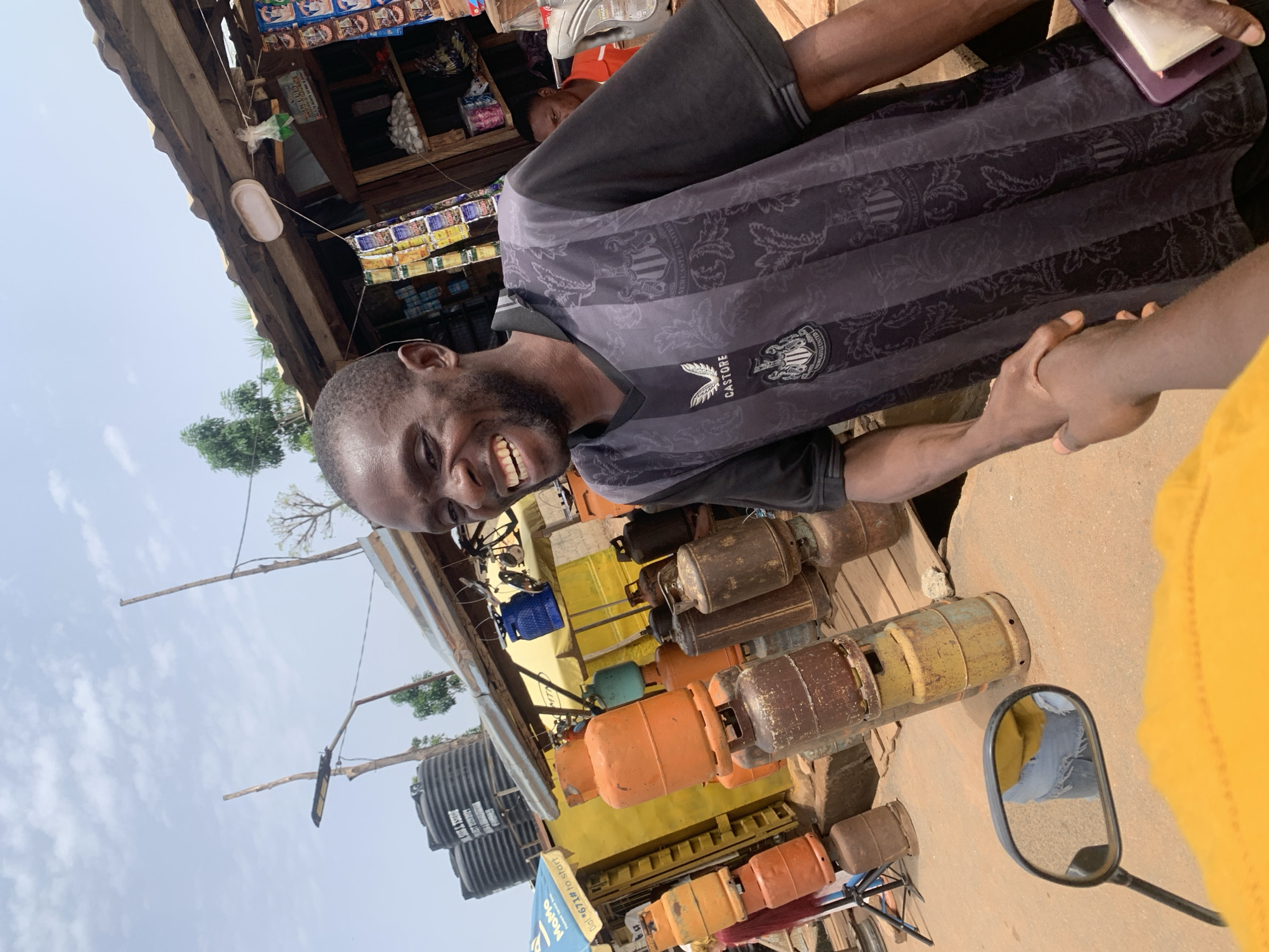 A man wearing a black and grey patterned Newcastle United jersey is smiling and looking directly at the camera. He is standing next to a display of various gas cylinders. The cylinders are in colors of orange, brown, yellow, and green. The background includes a building with a corrugated metal roof and a store with products visible on shelves. A water tank and street lights are also in the background. The scene appears to be taken from the perspective of someone on a motorbike, as a side mirror and part of a yellow garment are visible in the lower right corner of the image. This image was taken in Akwanga, Nigeria.