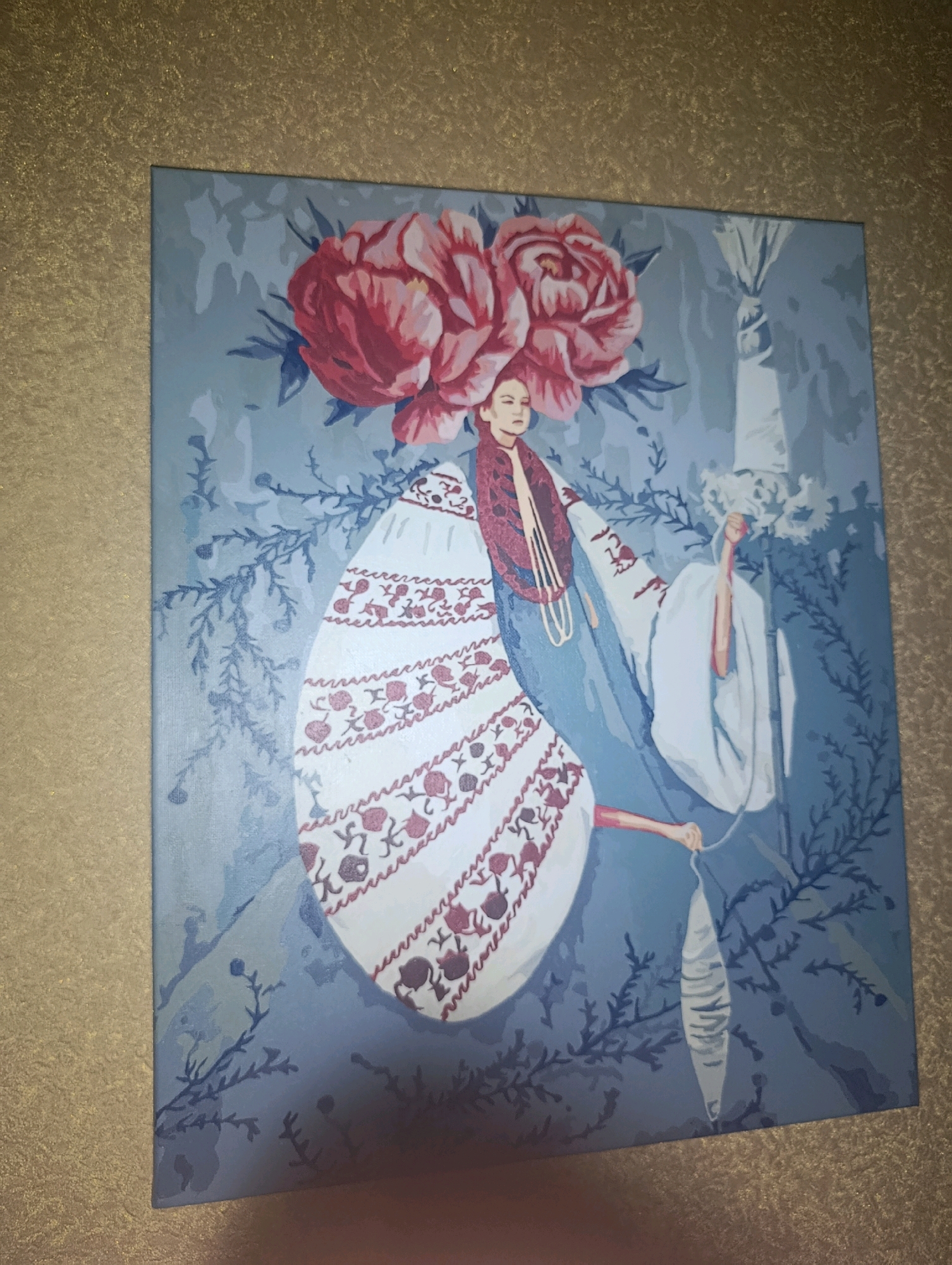 The image displays a painting featuring a woman adorned with large pink flowers on her head, resembling peonies. She is wearing a traditional costume characterized by a white dress with intricate red embroidery and a dark red outer garment. Her arms are extended, and she is holding a white object in her right hand. The background of the painting is a muted blue with a subtle floral or leafy pattern in a darker blue.
The painting is mounted on a textured, gold-colored surface, possibly a wall or a frame. There are no identifiable landmarks or elements within the painting that would suggest a specific location in the city of Horishni Plavni, Ukraine. The painting itself appears to be a piece of art, potentially a decorative item.