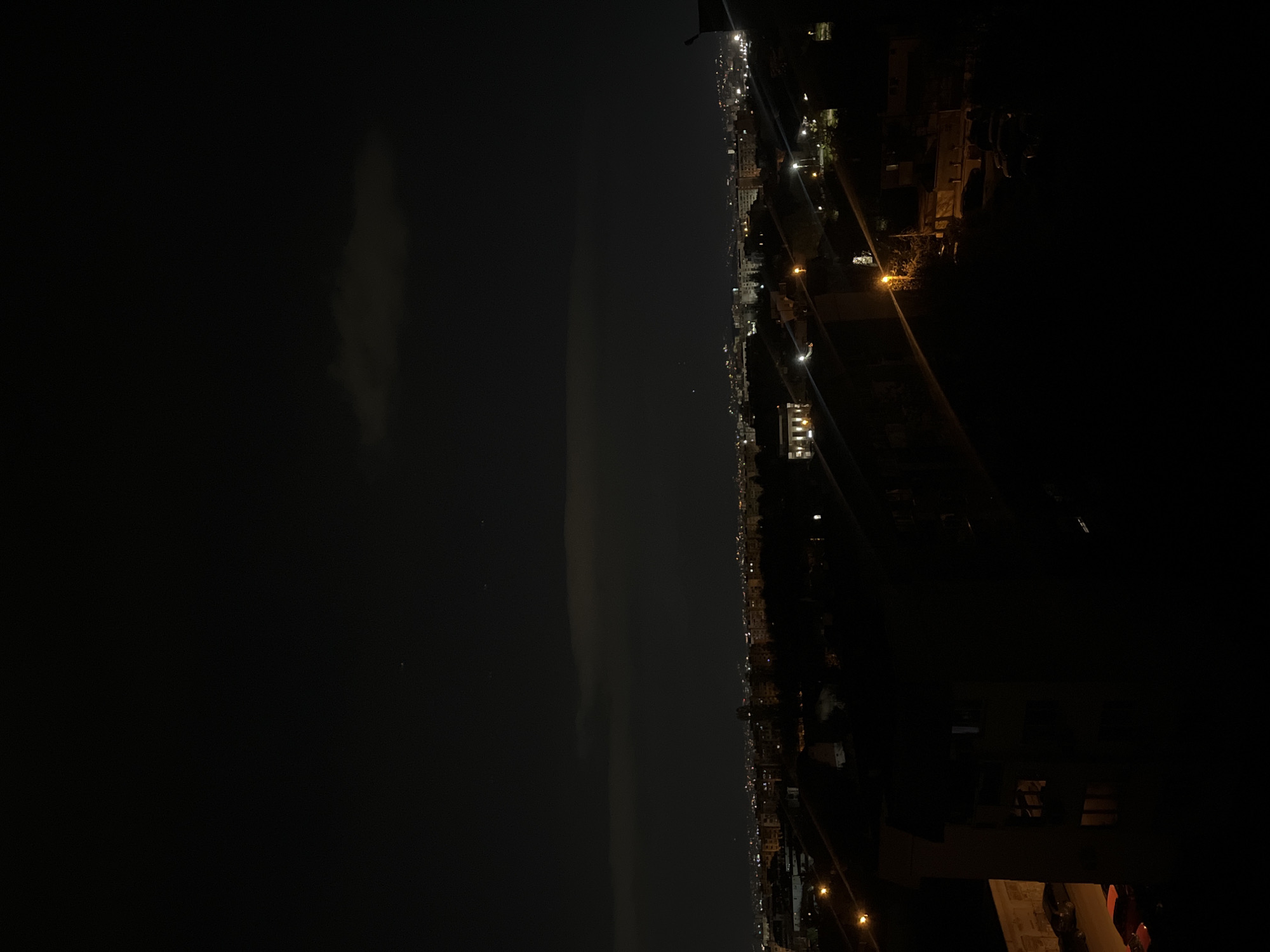 The image shows a nighttime cityscape, likely of Sofia, Bulgaria, based on the urban layout and architectural styles. The sky is mostly dark with some clouds. The right side of the image is dominated by the city, with buildings illuminated by streetlights and lights from inside buildings. On the left, the sky is dark with a visible cloud formation and some stars. The image appears to be taken from an elevated position, possibly a balcony or high floor of a building.