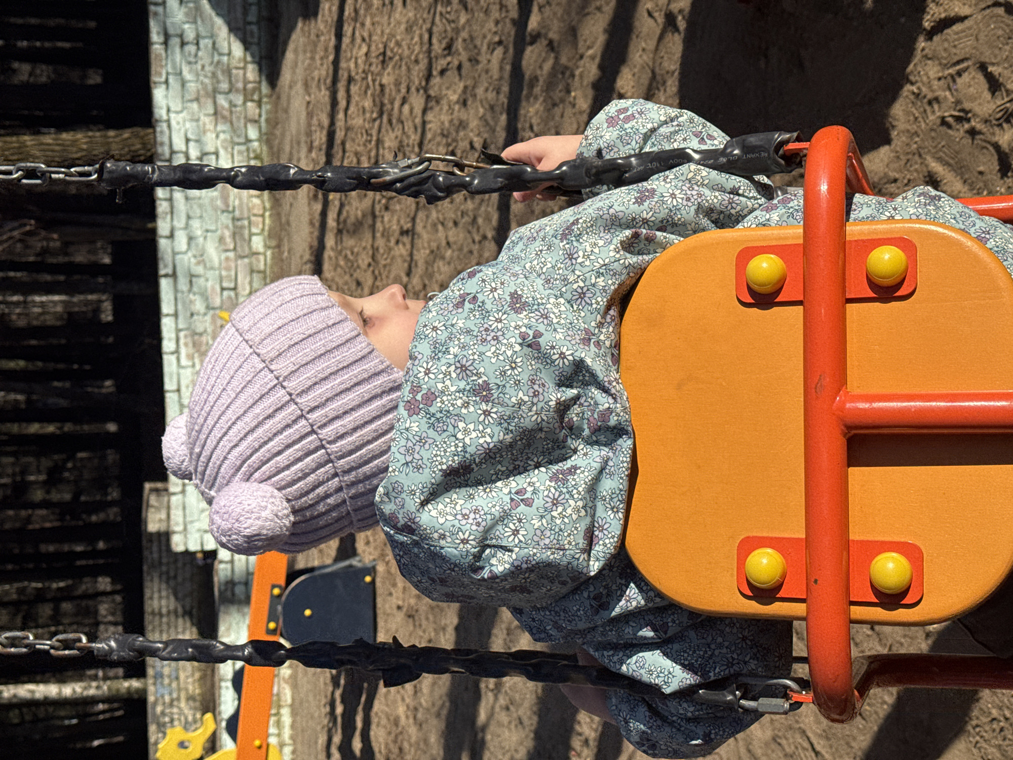 A young child, likely a girl, is captured sitting in a swing at an outdoor playground in Chaikovsky, Russia. The image is brightly lit by sunshine, casting strong shadows and indicating a clear daytime setting.
The child is seen from the chest up, wearing a light purple knitted beanie adorned with two fluffy pom-poms on top. She has a light blue long-sleeved dress with a delicate white and purple floral pattern. Her hands are grasping the black-covered chains of the swing, and her face is visible in profile, looking slightly upwards and to the left with a neutral expression.
The swing itself features a prominent orange seat with a red metal frame that forms a protective bar in front of the child. Two bright yellow spherical buttons or knobs are affixed to the top of the orange seat. The swing's suspension system consists of black, rubberized or plastic-coated chains, which appear to be protective coverings over metal chains. On one of these coverings, faint text can be discerned, reading "600V 110C" and "REXNANT GLOE."
The background of the playground shows textured ground, possibly sand or dirt, and various structures. To the left, a light-colored wall with a brick-like pattern is visible, alongside darker wooden elements. Strong shadows stretch across the ground, indicating the position of the sun and surrounding playground equipment.