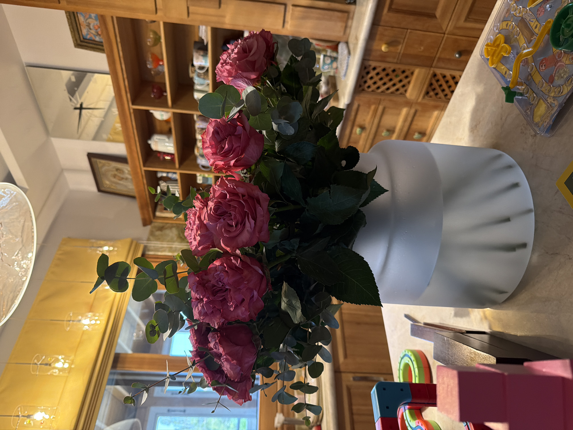 A bouquet of pink roses and eucalyptus leaves is displayed in a frosted glass vase on a marble countertop. Behind the vase, a wooden cabinet with multiple shelves is filled with various items, including mugs, decorative objects, and framed artwork. To the left of the vase are colorful building blocks and a toy. The background shows kitchen cabinetry and a window with yellow blinds. The scene is indoors, likely in a residential setting.
