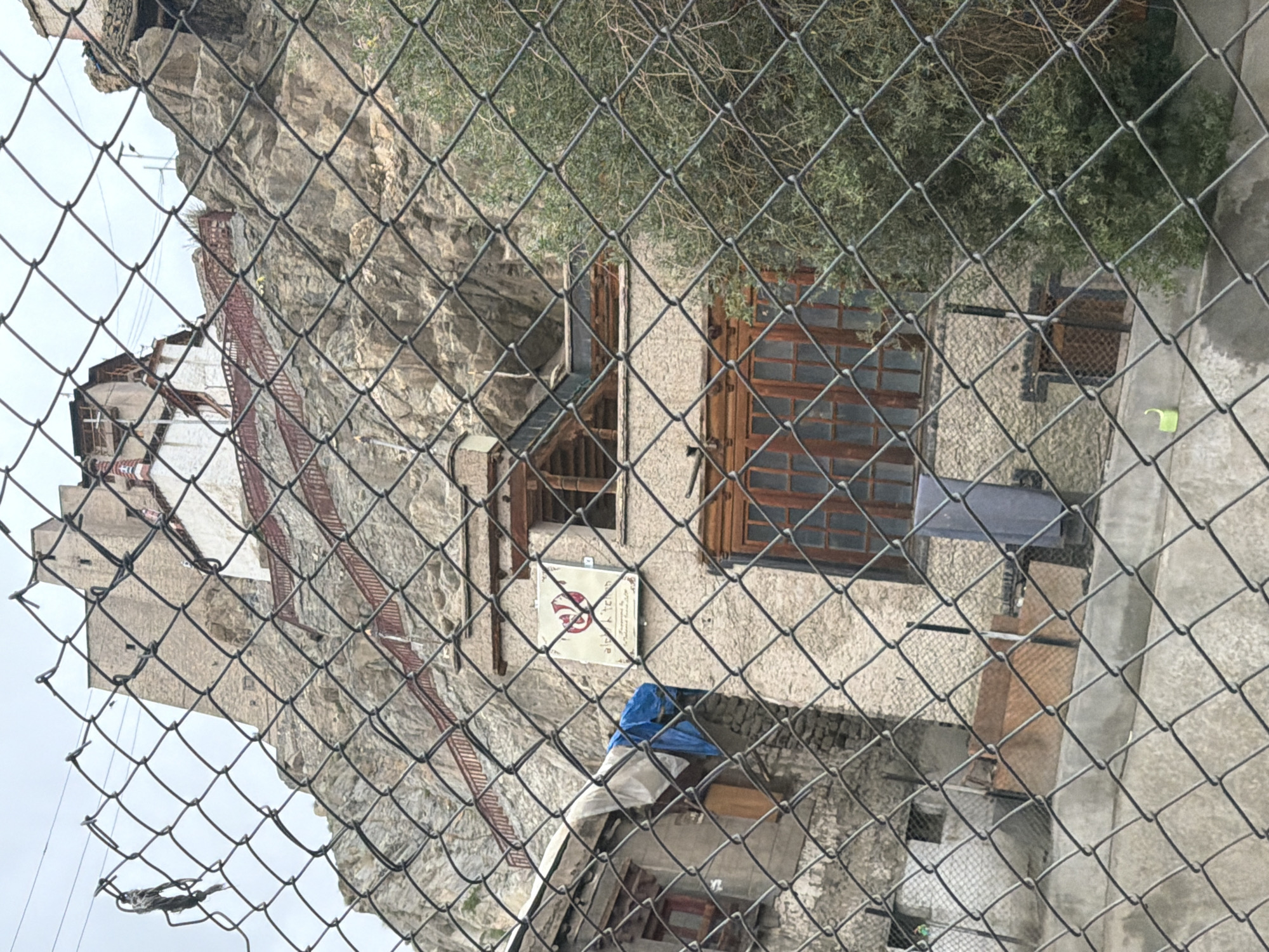 A chain-link fence is in the foreground, partially obscuring a view of a multi-story building built into a rocky hillside. The building has a textured, light-colored facade and wooden-framed windows. A sign with red lettering and a circular emblem is mounted on the building's wall. Red railings line a staircase ascending the hillside behind the building, leading to a larger structure at the top. The sky is overcast. The location is the Leh Palace in Leh, India.