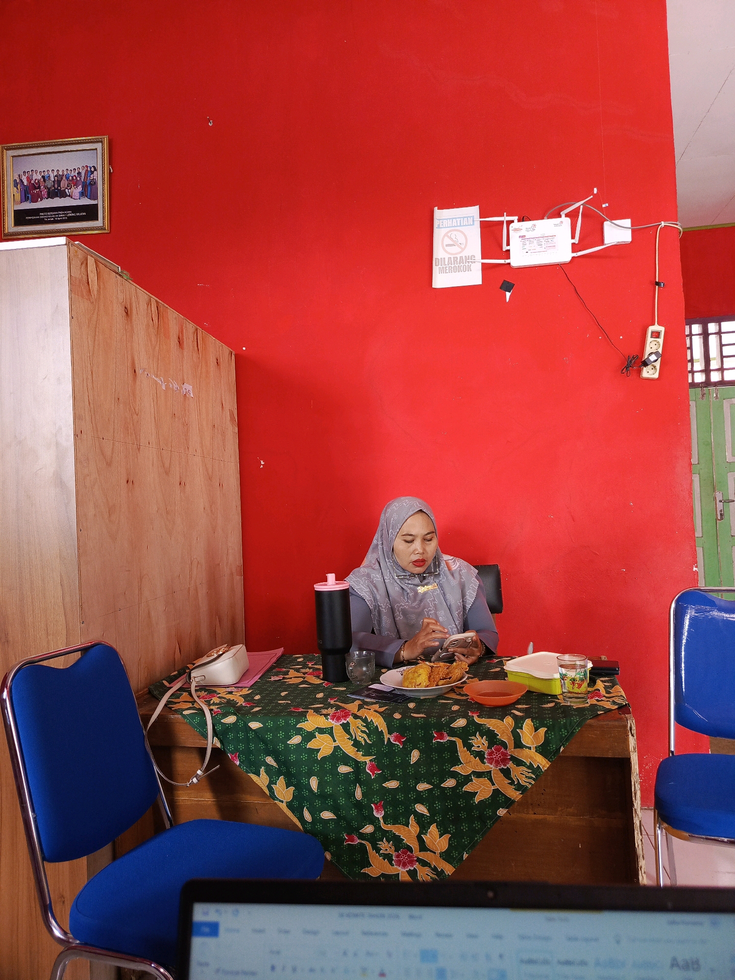 The photograph captures an indoor scene, likely within an office or a small establishment, with predominantly red walls. In the mid-ground, a woman wearing a grey hijab and a matching grey top with intricate patterns is seated at a table covered with a green and yellow batik-patterned cloth. She is holding a mobile phone in her left hand and appears to be eating from a plate in front of her. On the table, there are also a glass of water, a thermal flask, a small orange bowl, and a food container. Two blue chairs with metal frames are visible, one on the left and another on the right, partially framing the table. A wooden cabinet is positioned to the left of the woman, and a framed group photograph hangs on the red wall above it. On the upper right portion of the red wall, a white router with antennas and a white electrical outlet with a black cord are mounted. The environment suggests a functional space, possibly in Tik Jeniak, Indonesia, given the presence of Indonesian signage ("PERHATIAN DILARANG MEROKOK" which translates to "ATTENTION NO SMOKING"). In the foreground, a blurred laptop screen is visible, indicating a potential work-related setting.