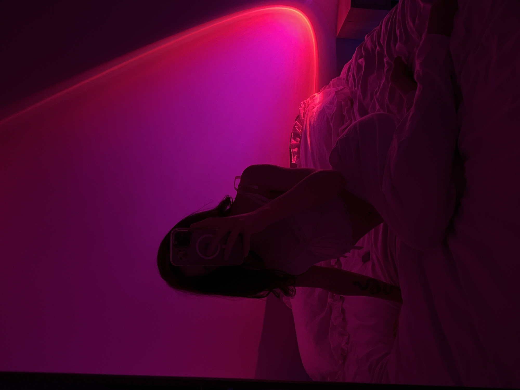 A person is taking a mirror selfie in a dimly lit bedroom. The person is seated on a bed, facing a mirror. They are wearing light-colored clothing. The room is illuminated by a pink and purple gradient light, possibly from a sunset lamp, casting a warm glow on the walls and the bed. The bed is covered with white sheets that appear rumpled. A phone is held up to take the photo, obscuring the person's face. The background shows the wall with the distinctive gradient light pattern. There is a glimpse of a bedside table on the right side of the image. The scene is intimate and focuses on the aesthetic created by the lighting.