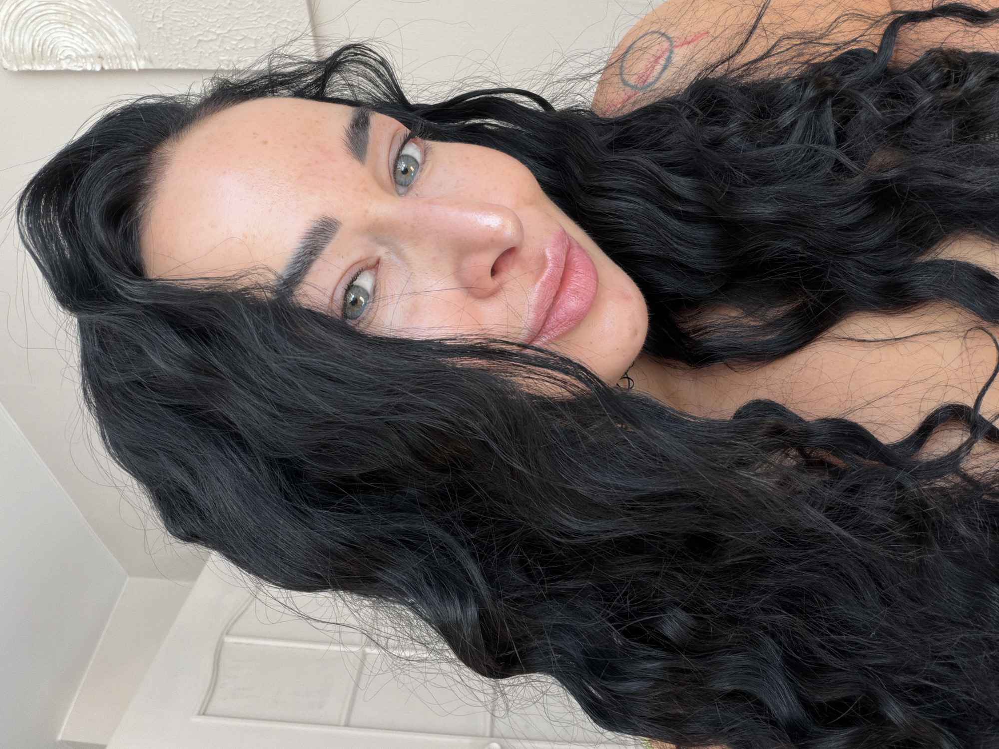 A close-up portrait shows a woman with fair skin, dark, wavy hair, and light-colored eyes. She is looking slightly up at the camera. She has noticeable freckles, particularly on her nose and cheeks. Her lips have a glossy appearance. Part of a tattoo is visible on her shoulder. The background includes a white textured wall. The location is Unknown, Unknown.