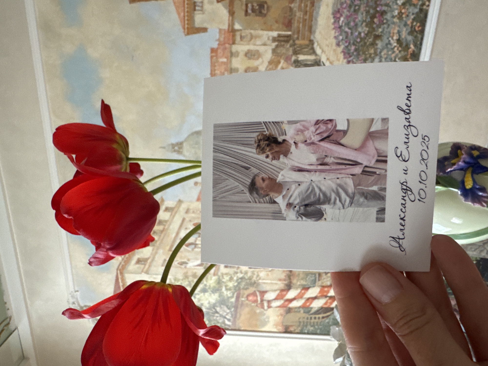 A photograph shows a white card, possibly a wedding invitation, being held in a person's hand with visible polished fingernails. The card features a black-and-white photograph of a couple, likely the wedding couple. The invitation has black cursive script that reads "Александр и Елизавета 10.10.2025" which translates to Alexander and Elizabeth, 10/10/2025, written on it. Red tulips are arranged nearby the invitation, slightly out of focus, suggesting they are in the foreground. In the background is a blurry painting, which could be a mural, showing architectural elements reminiscent of Saint Petersburg, Russia, featuring buildings, trees, and a cityscape.