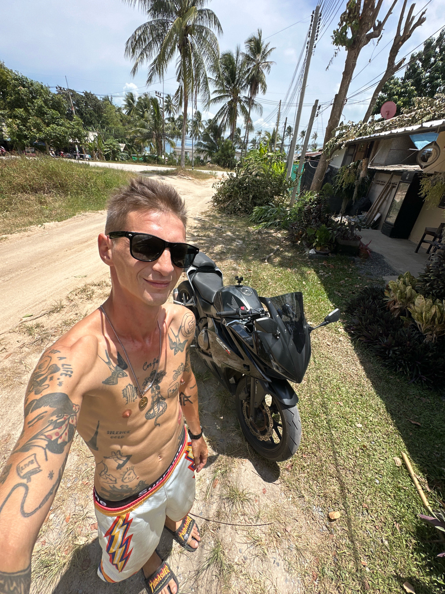 A man with extensive tattoos on his torso and arms is taking a selfie outdoors. He is wearing sunglasses and white shorts with a lightning bolt pattern. Next to him is a black motorcycle. The background shows a dirt road, palm trees, and a simple building with an antenna. The scene appears to be in a tropical, possibly rural, location on the island of Koh Phangan, Thailand, given the vegetation and style of building. The lighting suggests it is daytime with bright sunlight.
