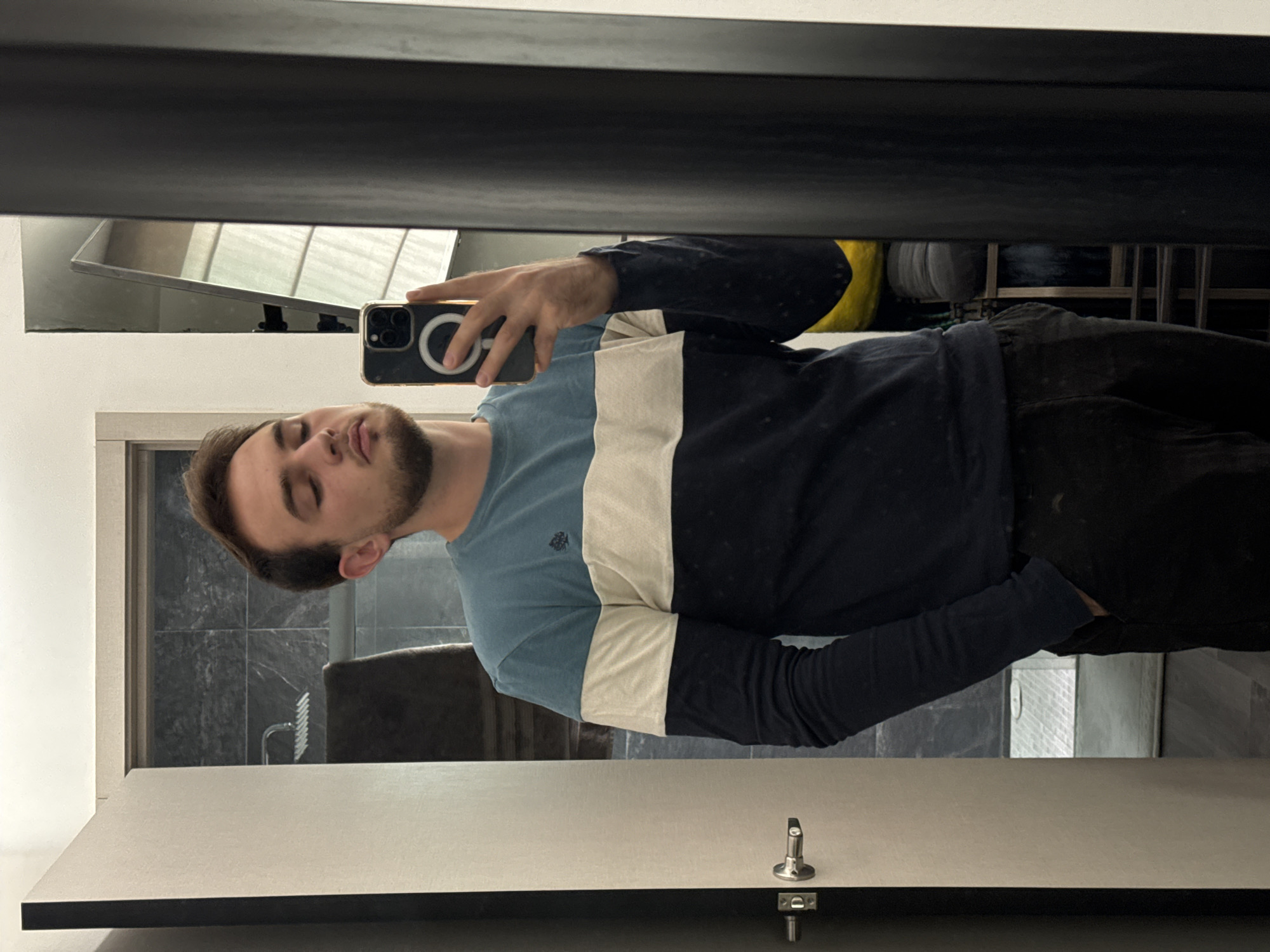 A man is taking a selfie in a bathroom. He is wearing a blue, white, and navy blue long-sleeved shirt and black pants. He is holding a black smartphone with a white ring on the back. Behind him is a bathroom stall door and a sink with a mirror. The bathroom stall has a black frame. There are grey tiles on the wall. The toilet paper holder is visible. It is not possible to pinpoint the location in the city of San Bernardino Tlaxcalancingo, Mexico, from the image alone.