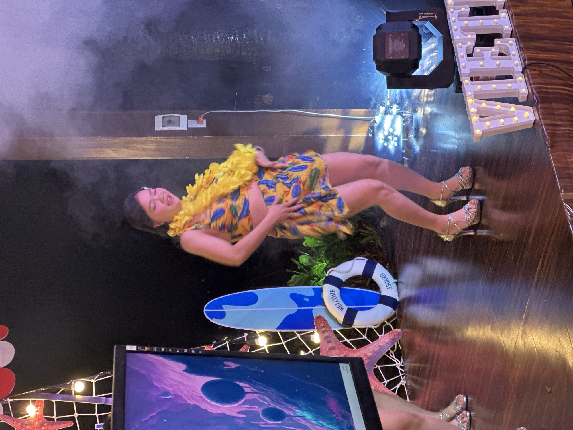 A woman wearing a yellow lei and a short, patterned dress is lying on a stage or platform decorated with beach-themed props. She's smiling, and her arms are positioned on her stomach.
Props include a surfboard, a life preserver with "Welcome Aboard" printed on it, and starfish on netting. A monitor or screen is in the foreground.
Overhead, stage lighting and fog contribute to the scene's ambience. A sign with illuminated letters is visible to the right. The location is Phu Thuy Ward, Vietnam.