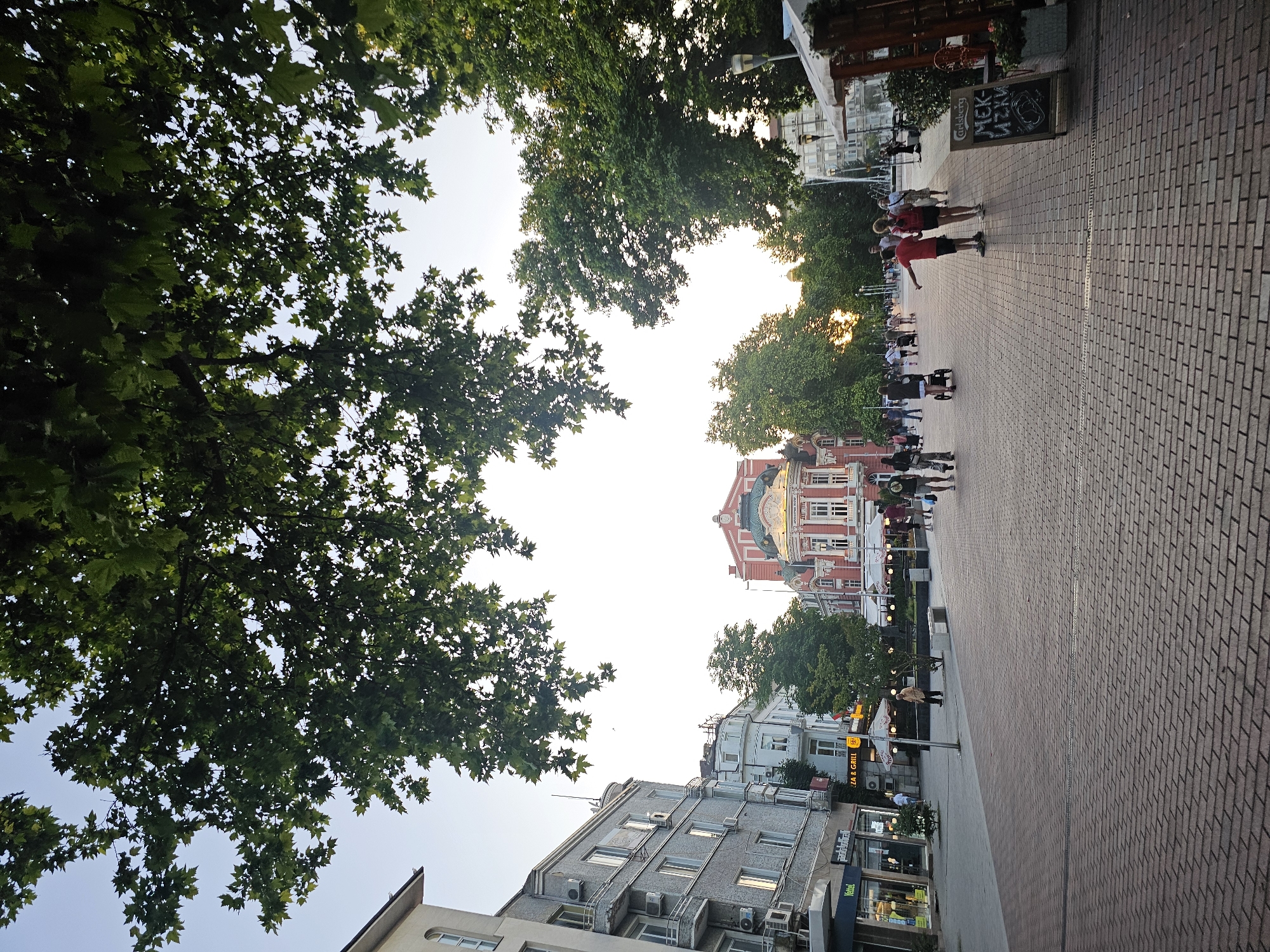 The image depicts a pedestrian street scene in an urban environment, likely in a European city given the architectural styles and the presence of a theater. A wide, brick-paved walkway is lined with buildings and trees. In the foreground, the brick pattern is clearly visible, with a slight tilt to the right. On the left, a multi-story building with grey facade and balconies, featuring numerous air conditioning units, dominates the view. Next to it, another building with lighter facade and visible shop fronts. Further down the street, a prominent, ornate building with red and cream coloring, featuring a distinctive gold dome and arched windows, stands out, appearing to be a theater or a similar cultural establishment. Several trees, some with large leaves, provide shade over the walkway. A variety of people are walking along the street, some in groups, others individually. Some appear to be tourists, with one person pointing towards the theater. There are also outdoor seating areas with umbrellas and a chalkboard sign indicating a menu. The lighting suggests it is late afternoon or early evening, with a soft glow from the setting sun visible through the trees in the distance. The overall atmosphere is one of casual urban activity.