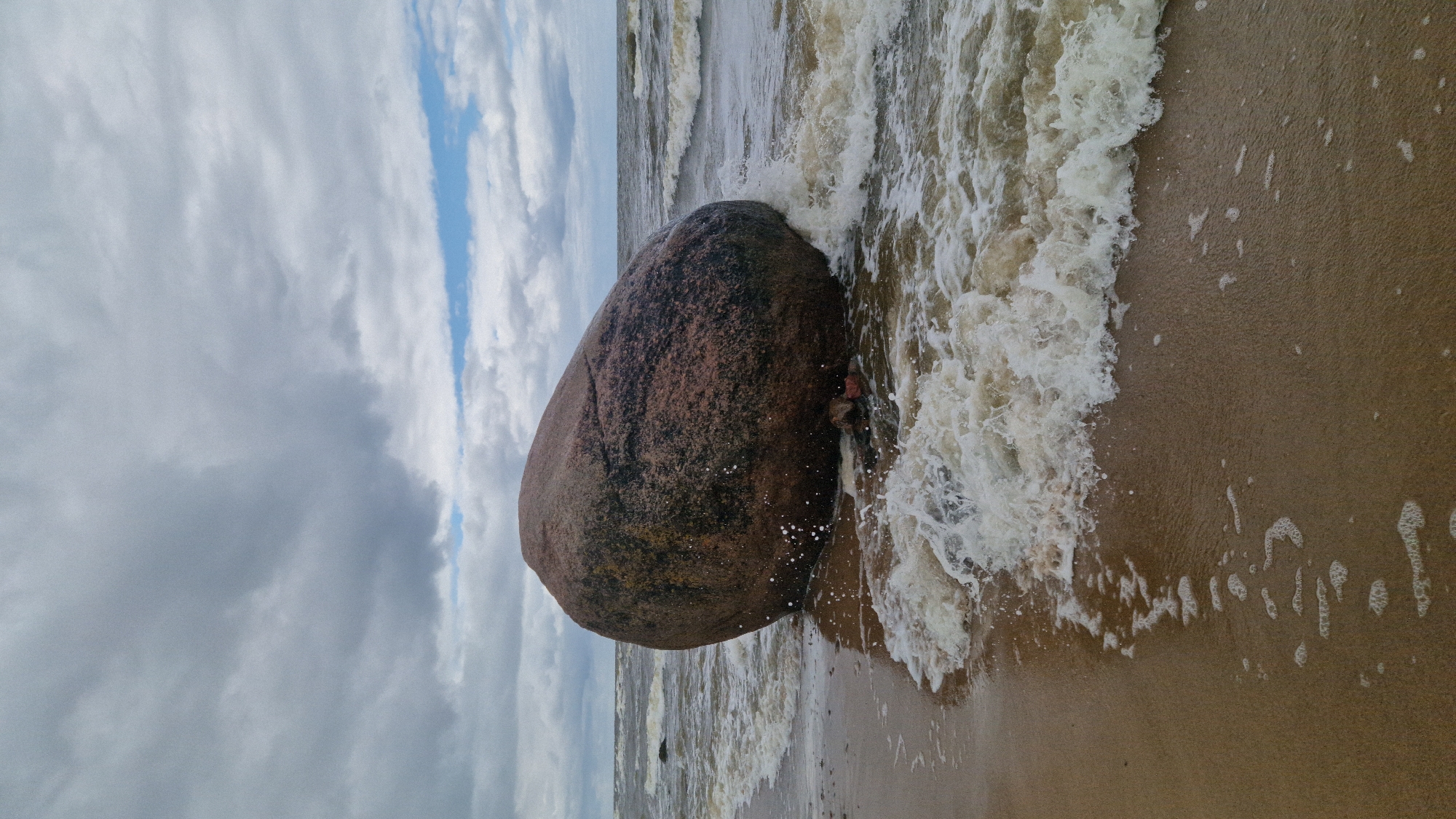 The image captures a large, dark boulder partially submerged in the choppy waters of the Baltic Sea on a cloudy day at Vārzas beach, Latvia. The boulder, rough and textured, is a dominant element in the foreground, with waves crashing against its base and creating white foam. The wet sand in the foreground shows receding water patterns and small bubbles. The sea extends to the horizon, meeting a sky filled with dramatic, grey clouds, with occasional patches of blue visible. The overall atmosphere is one of raw, natural beauty and the power of the sea. No people are present in the scene.