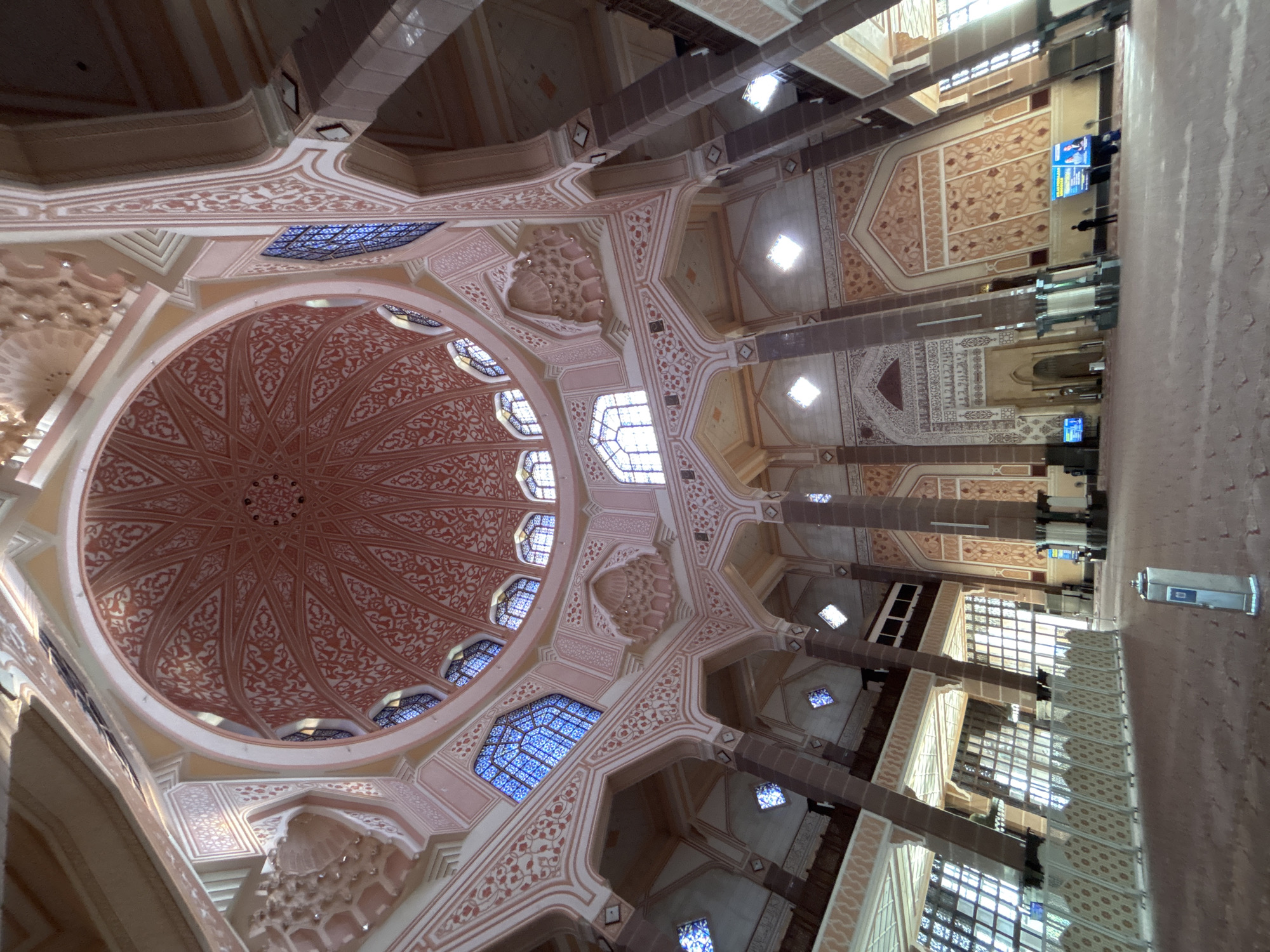 The image shows the interior of the Putra Mosque in Putrajaya, Malaysia, looking upwards towards the grand dome. The architecture features intricate pink and white decorative patterns on the dome and surrounding arches, supported by tall, dark columns. Blue stained-glass windows are visible along the upper walls. The floor is covered in a light-colored carpet, and a low, decorative barrier runs along the left side of the image. Several people are present, mostly standing or walking in the distance. Digital screens are mounted on some of the pillars. Sunlight streams in through large windows on the left side of the mosque.