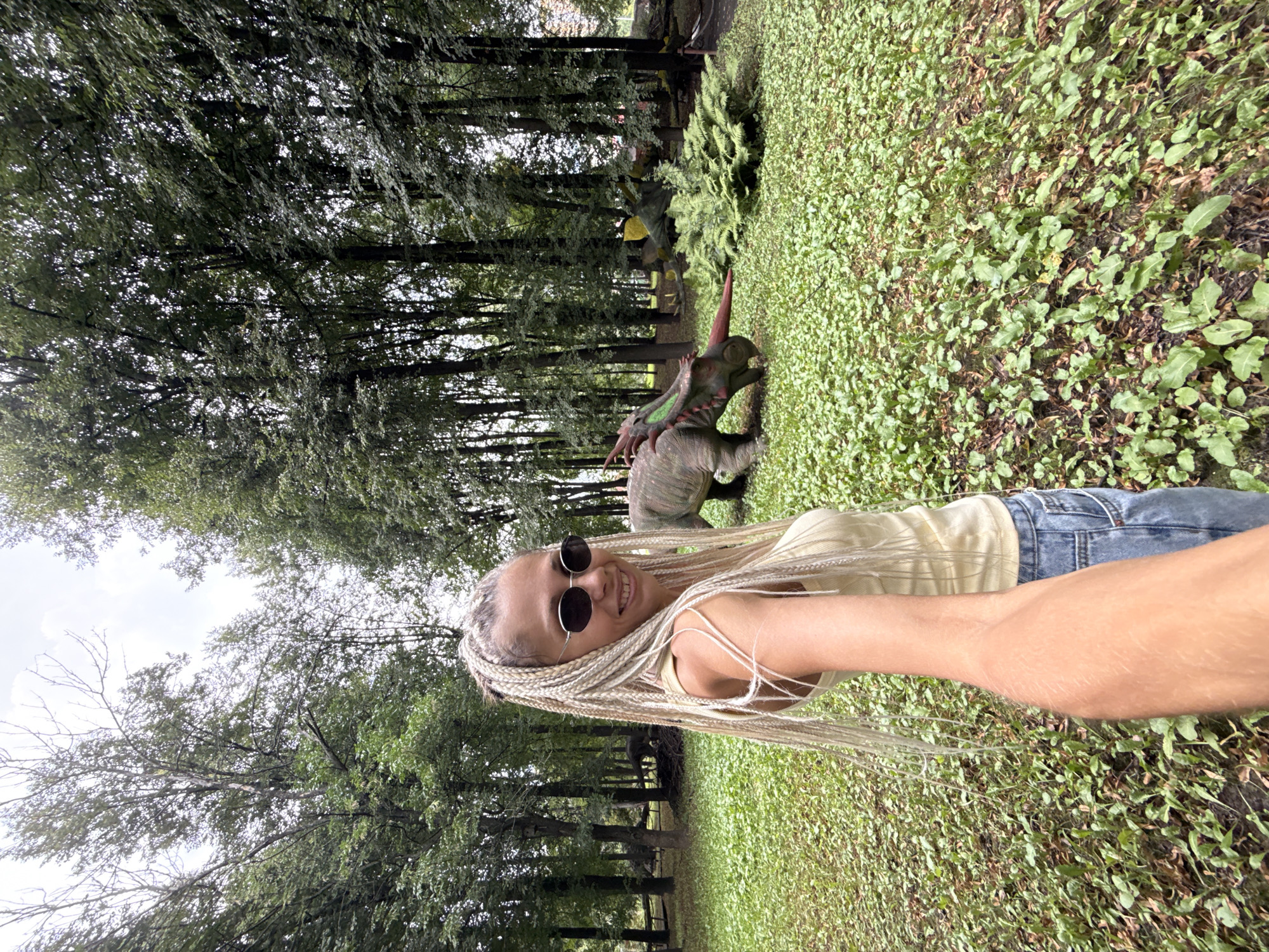 In Rusanovo, Russia, a woman with blonde braided hair and sunglasses takes a selfie in a park. She is wearing a light-colored tank top and denim jeans. In the background, several dinosaur sculptures are displayed amongst lush green trees and ground cover. One sculpture, a Triceratops, is prominently featured in the mid-ground, with another dinosaur partially visible behind it. The scene appears to be an outdoor exhibit within a wooded area.