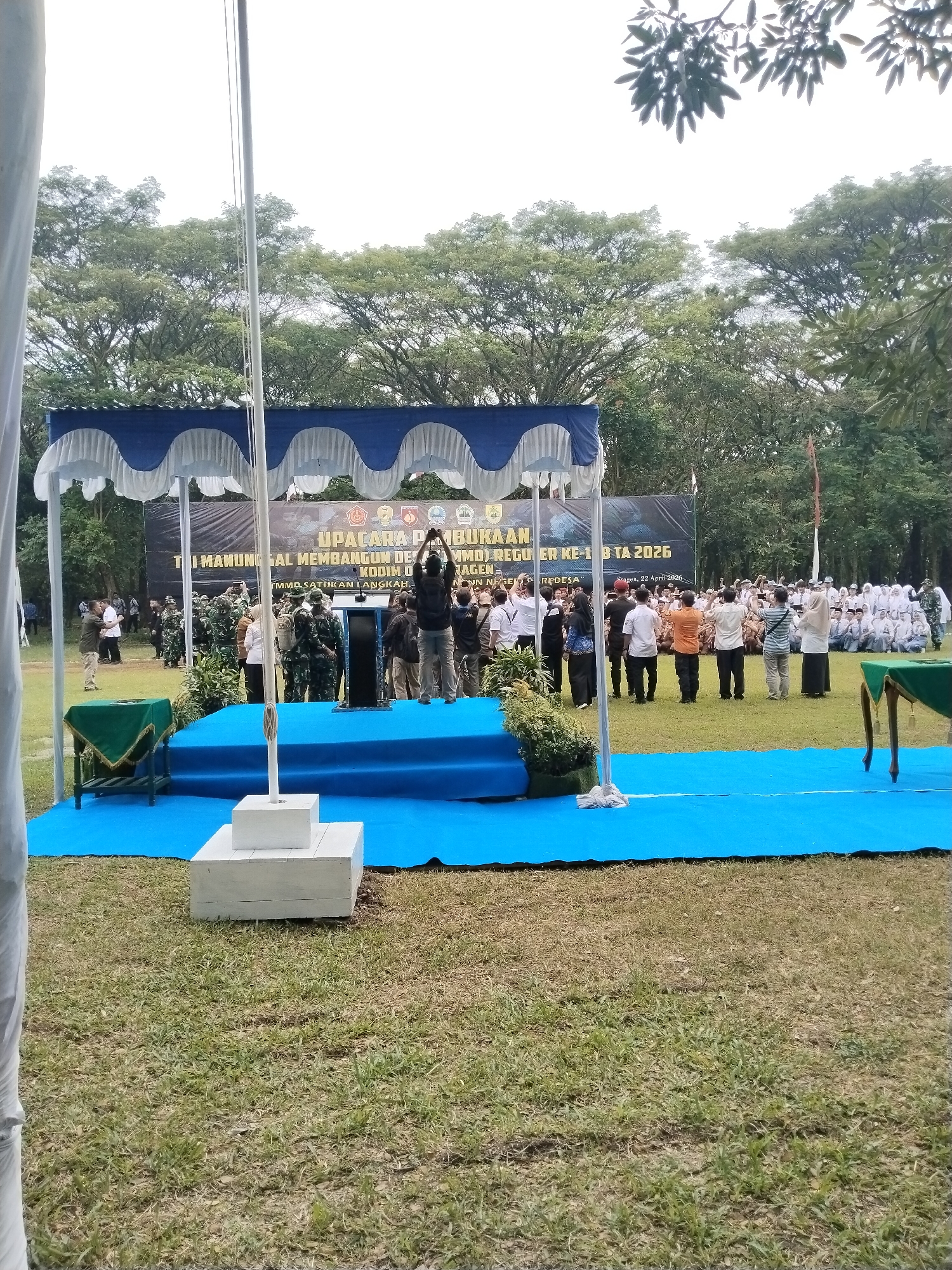The image depicts an outdoor opening ceremony in Puro, Indonesia, held on a grassy field surrounded by tall trees under an overcast sky. A central raised platform, covered in blue carpeting, is sheltered by a blue and white striped canopy. A flag pole stands prominently on the platform.
Behind the platform, a large banner with text and logos serves as a backdrop. Visible text includes "UPACARA PEMBUKAAN" (Opening Ceremony), "TNI MANUNGGAL MEMBANGUN DESA (TMMD) REGULER KE-1 B TA 2026," "KODIM," and "TMMD SATUKAN LANGKAH." The banner also indicates "Sragen, 22 April 2026."
On the platform, several individuals are gathered, including military personnel in camouflage uniforms and civilians. One person is seen holding a ceremonial object. A group of onlookers stands in front of the platform, some actively taking photos or videos with their phones. Further in the background, a larger assembly of people, many dressed in white, are seated or kneeling on the grass. Two small tables draped in green cloth are positioned on the grass to the sides of the platform. The overall scene suggests an official military-civilian community development program launch or similar public event. The attendees appear attentive and engaged in the ceremony.
