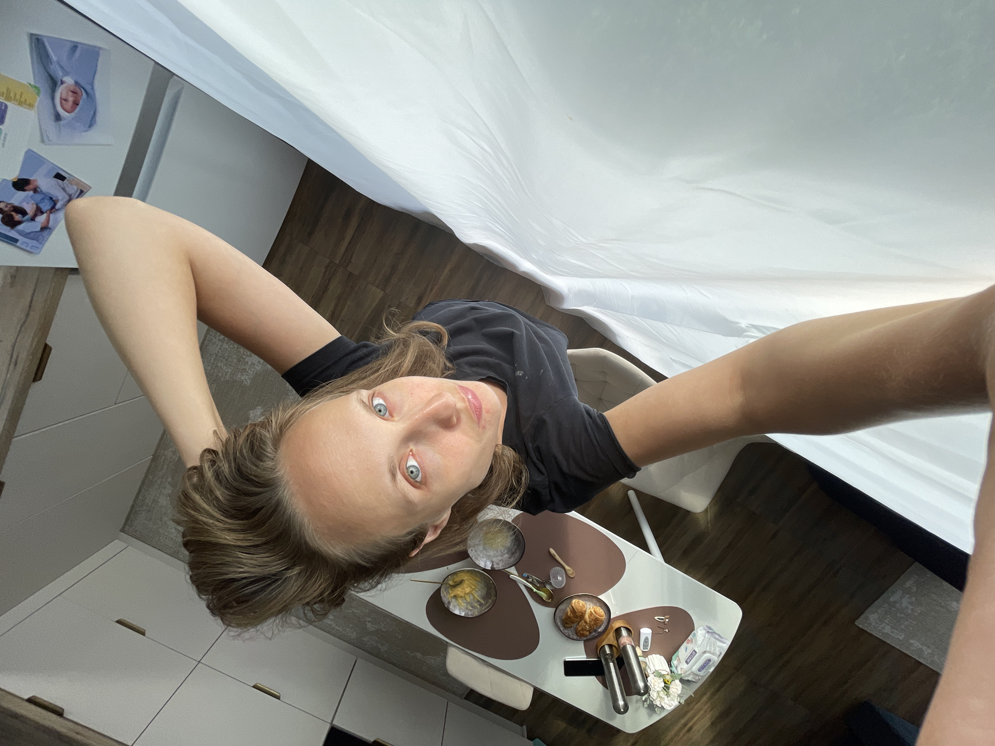 A high-angle, wide shot captures a woman in a black t-shirt holding her right arm up with her left hand in her hair, looking upwards at the camera. She has blue eyes and brown hair styled in a high ponytail. She is positioned in a kitchen, with a dining table in the foreground containing a smartphone, croissants, and various other items. In the background, there are white cabinets and a refrigerator adorned with photographs. The floor is made of wood. The overall scene suggests a domestic environment within the city of Podshivalovo, Russia.