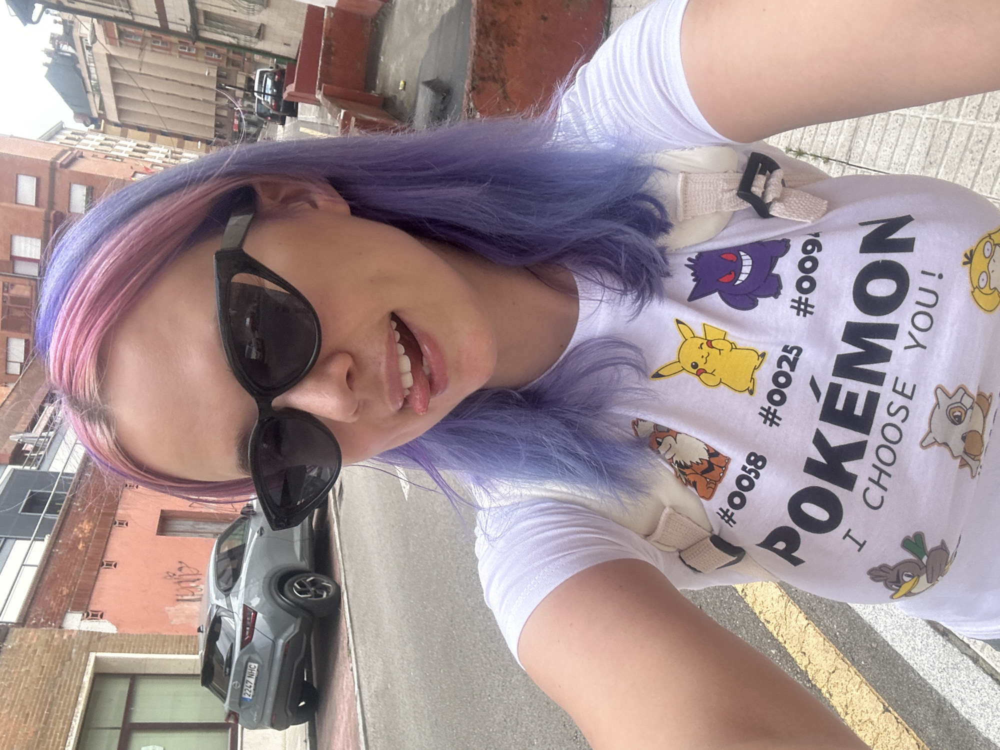 A young woman with pink and purple dyed hair, wearing black cat-eye sunglasses, a white t-shirt with Pokemon characters and the text "POKÉMON I CHOOSE YOU!", and a light-colored backpack, sticks her tongue out playfully for a selfie. She is standing on a sidewalk in Oviedo, Spain, with a grey Nissan Juke parked on the street to her left. Buildings with reddish-brown and beige facades line the street behind her. The image captures an urban street scene in Oviedo, with a focus on the woman and her distinctive style.