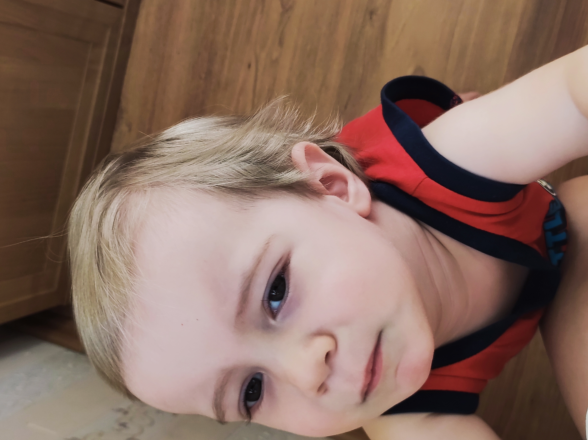 A close-up, slightly angled portrait captures a young child with fair skin, blonde hair, and dark eyes. The child is wearing a red shirt with a navy blue neckline. The background consists of wooden flooring and a cabinet. The location is implied to be Chaikovsky, Russia, based on the instructions.