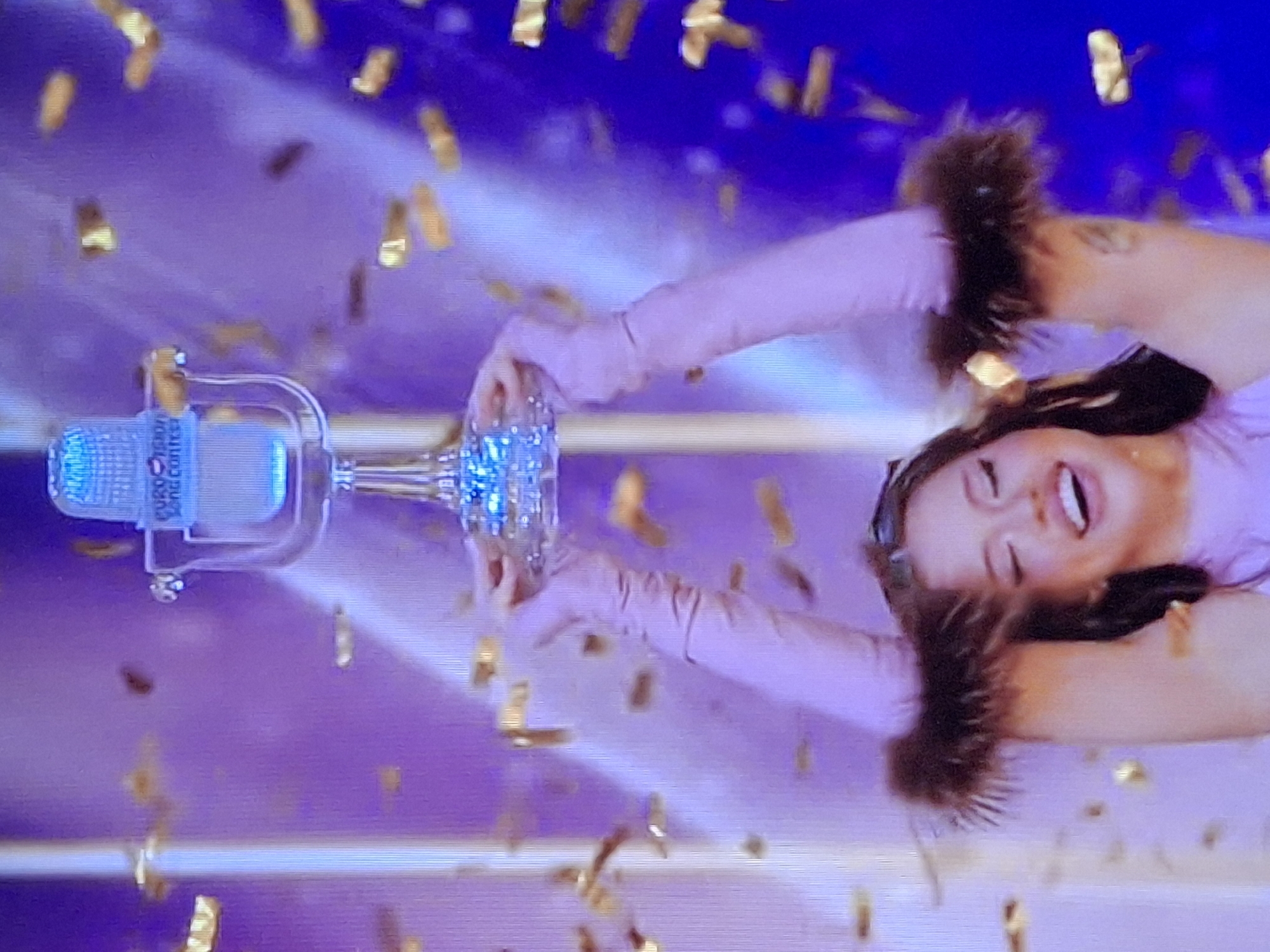 The image shows a woman holding what appears to be a Eurovision Song Contest trophy. She is smiling and wearing long pink gloves with fur cuffs. Gold confetti is falling around her. The background is a blur of purple and blue light. Location: Unknown, Unknown.