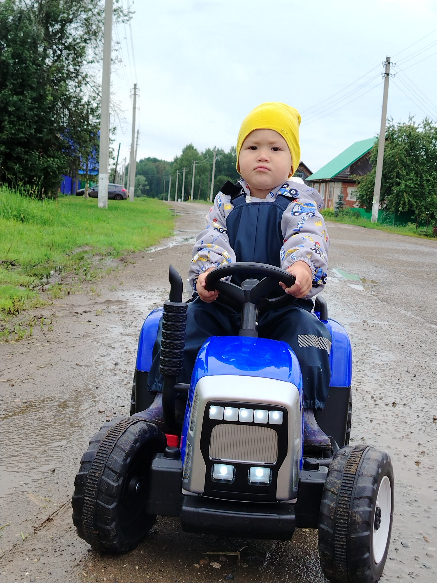 A young child wearing a yellow hat and blue overalls is sitting in a blue toy tractor outdoors. The child is holding the steering wheel and looking at the camera. The tractor is on a wet, dirt road. In the background, there are trees, power lines, and houses. The scene appears to be in a rural area, possibly the city of Gorzhan, Russia.