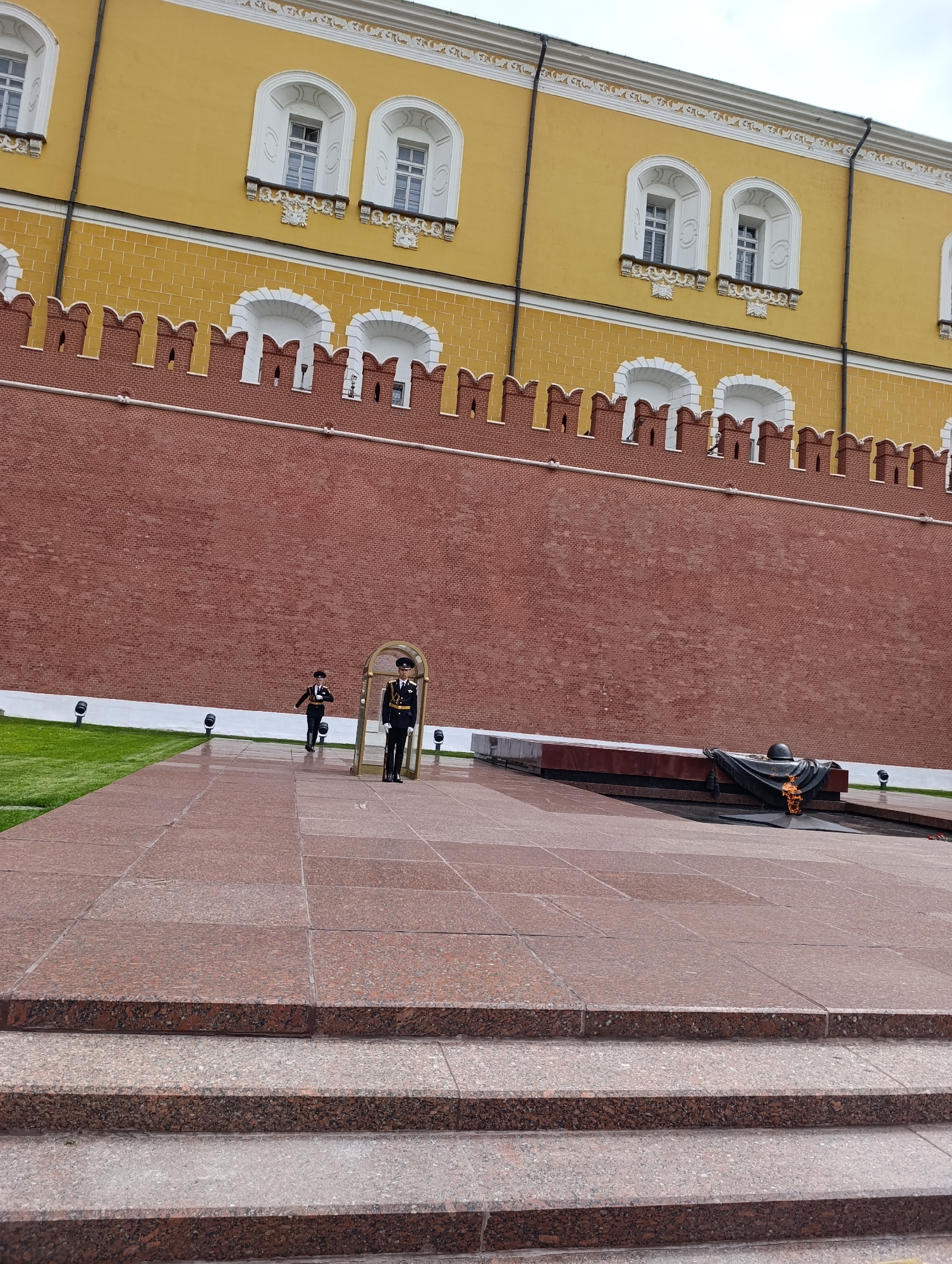 The image captures an outdoor ceremonial scene in front of a prominent historical wall. In the foreground, several steps made of speckled reddish-brown granite lead up to a large paved area of similar material. To the right of this paved area, a dark, low-lying memorial structure is visible. It features a five-pointed star with a perpetual flame, a military helmet, and a dark fabric drape.
Two individuals in dark military uniforms are present. One stands at attention within an ornate golden archway to the right, near the memorial. The second uniformed person is walking from the left side of the frame towards this archway, indicating a possible guard change or ceremonial patrol.
Behind the paved area and the memorial, a narrow strip of well-maintained green grass is visible, illuminated by small ground-level lights. The backdrop is dominated by a tall, red brick wall with crenelated battlements along its top edge. Above this wall, a light yellow building rises, featuring multiple white arched windows with decorative sills and white trim. The sky appears overcast, and the ground shows signs of dampness, possibly from recent rain. No visible text is present in the image.