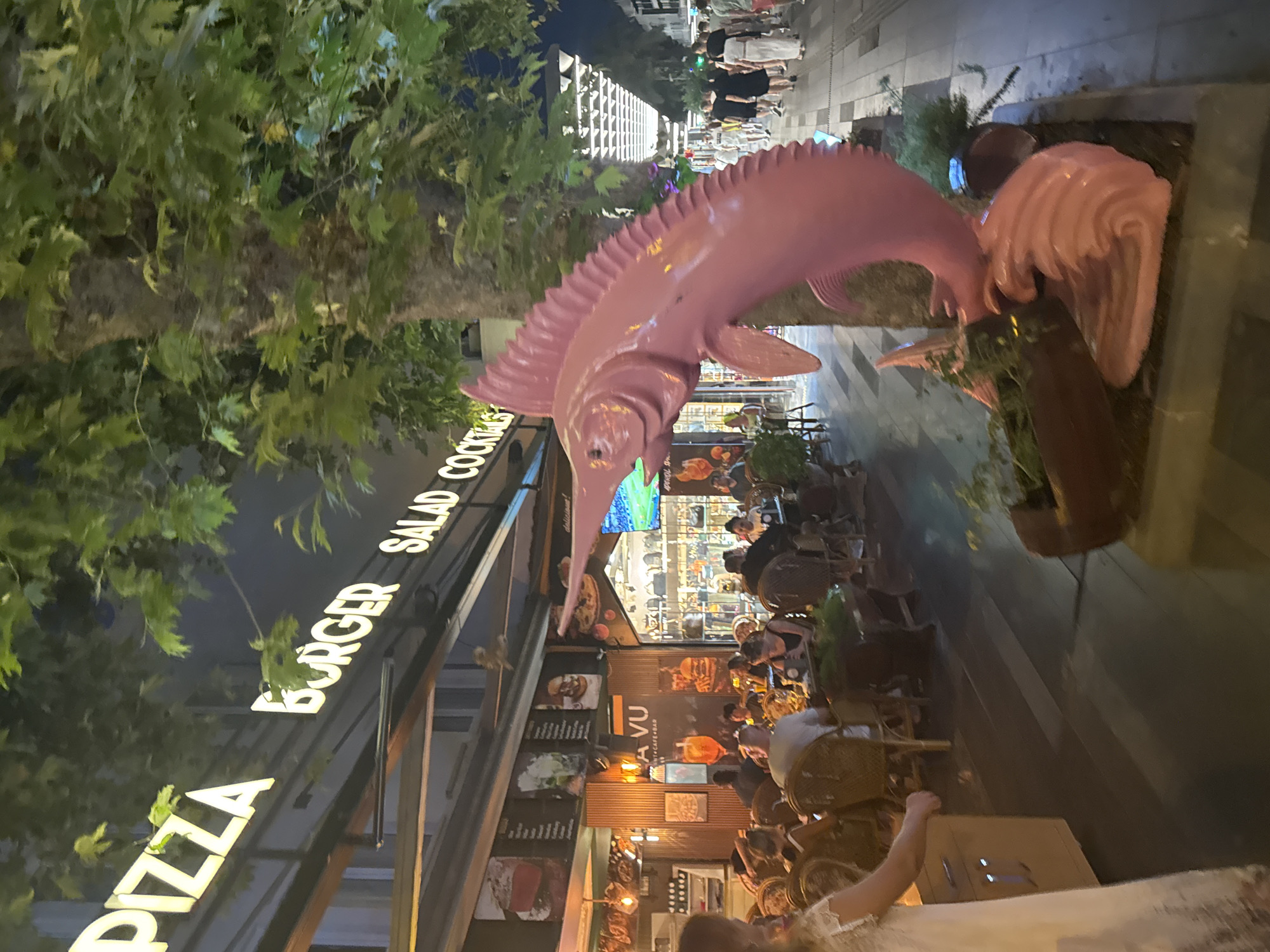 In Kemer, Turkey, this nighttime photograph captures a lively outdoor dining scene on a pedestrian street. A large, bright pink statue of a marlin, seemingly made of plastic or fiberglass, stands prominently in the foreground, leaning against a tree. Behind the statue, a restaurant with a lit facade features illuminated signage that reads "PIZZA BURGER SALAD COCKTAILS." Diners are seated at tables on a wooden deck, with some individuals visible in the background walking along the street, which is lined with shops and illuminated by overhead lights. The overall atmosphere suggests a bustling tourist area.